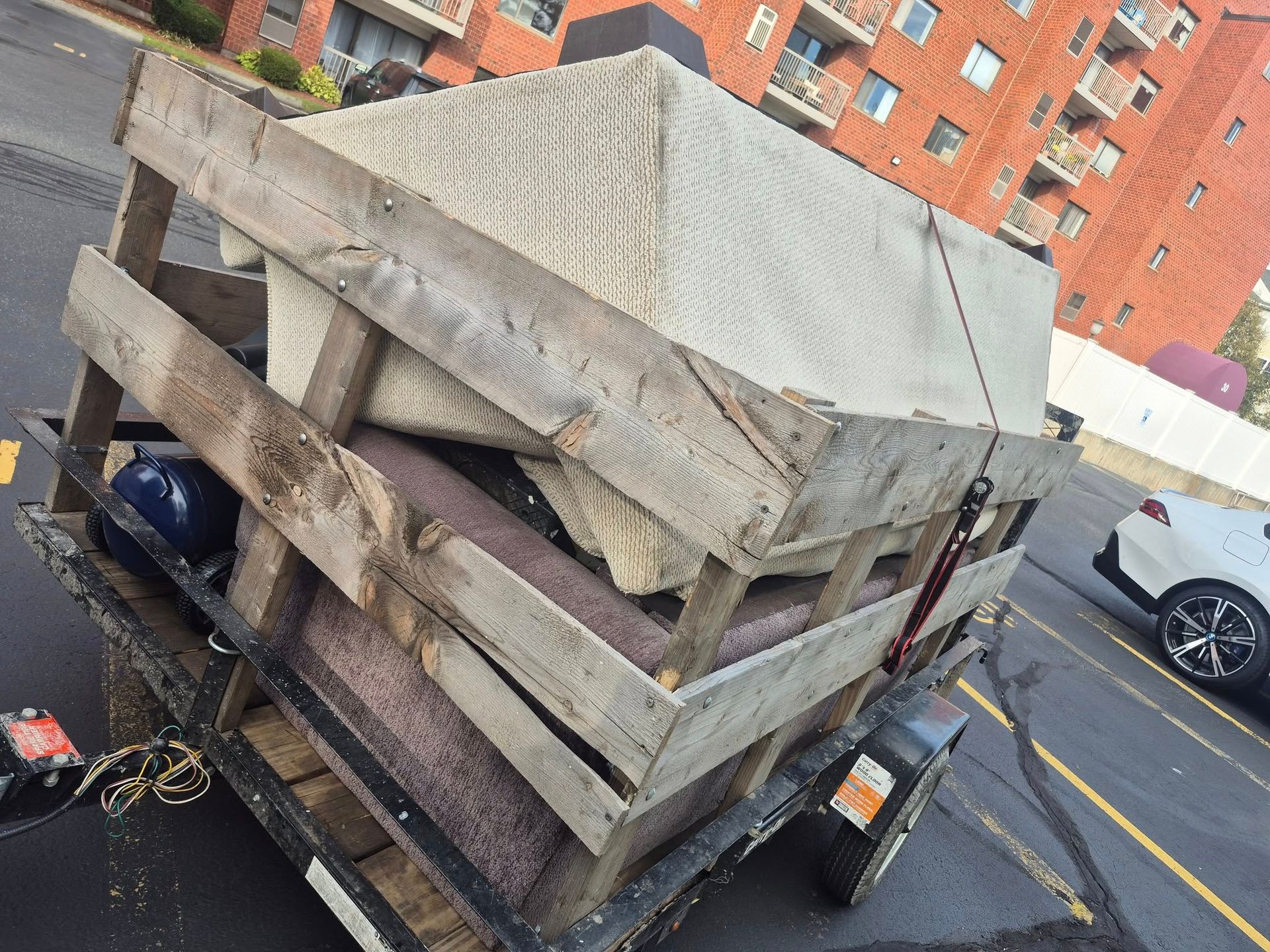 Trailer loaded with a mattress, carpet, and other items. Parked outside a brick apartment building.