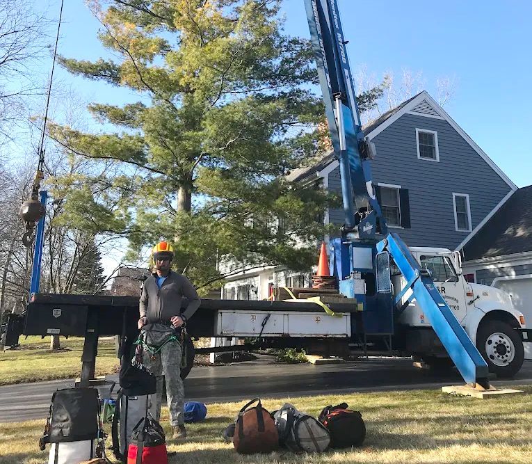 A worker in a helmet and harness stands next to a blue boom truck parked on a driveway in front of a house.