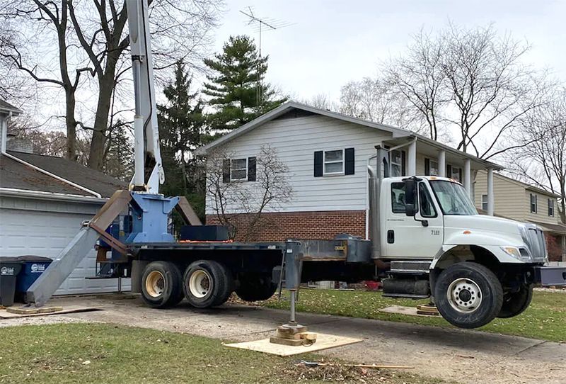 A white utility truck with a raised boom arm, stabilized by outriggers on a residential driveway.