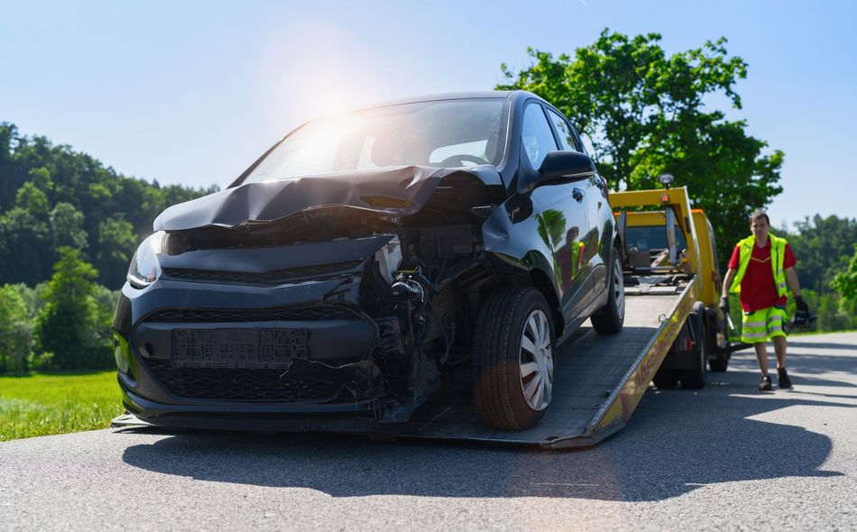 Damaged black car on a tow truck, roadside with a tow truck operator in a red vest and green shorts.