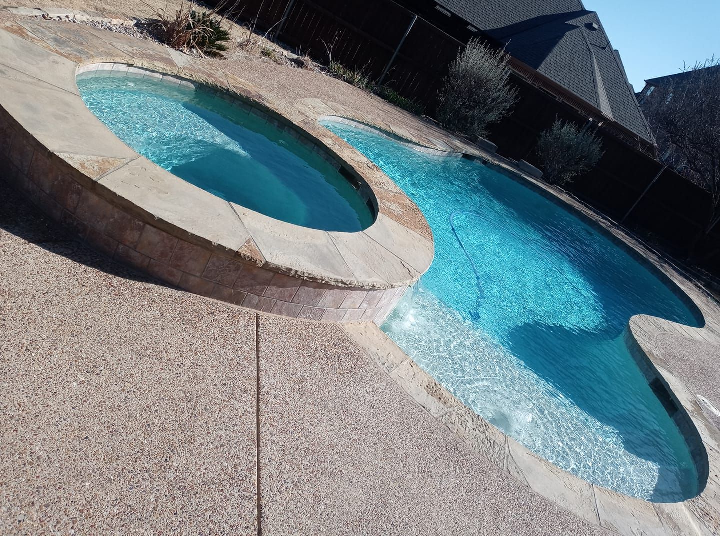 Swimming pool with attached hot tub, light blue water, beige stone border, residential setting.
