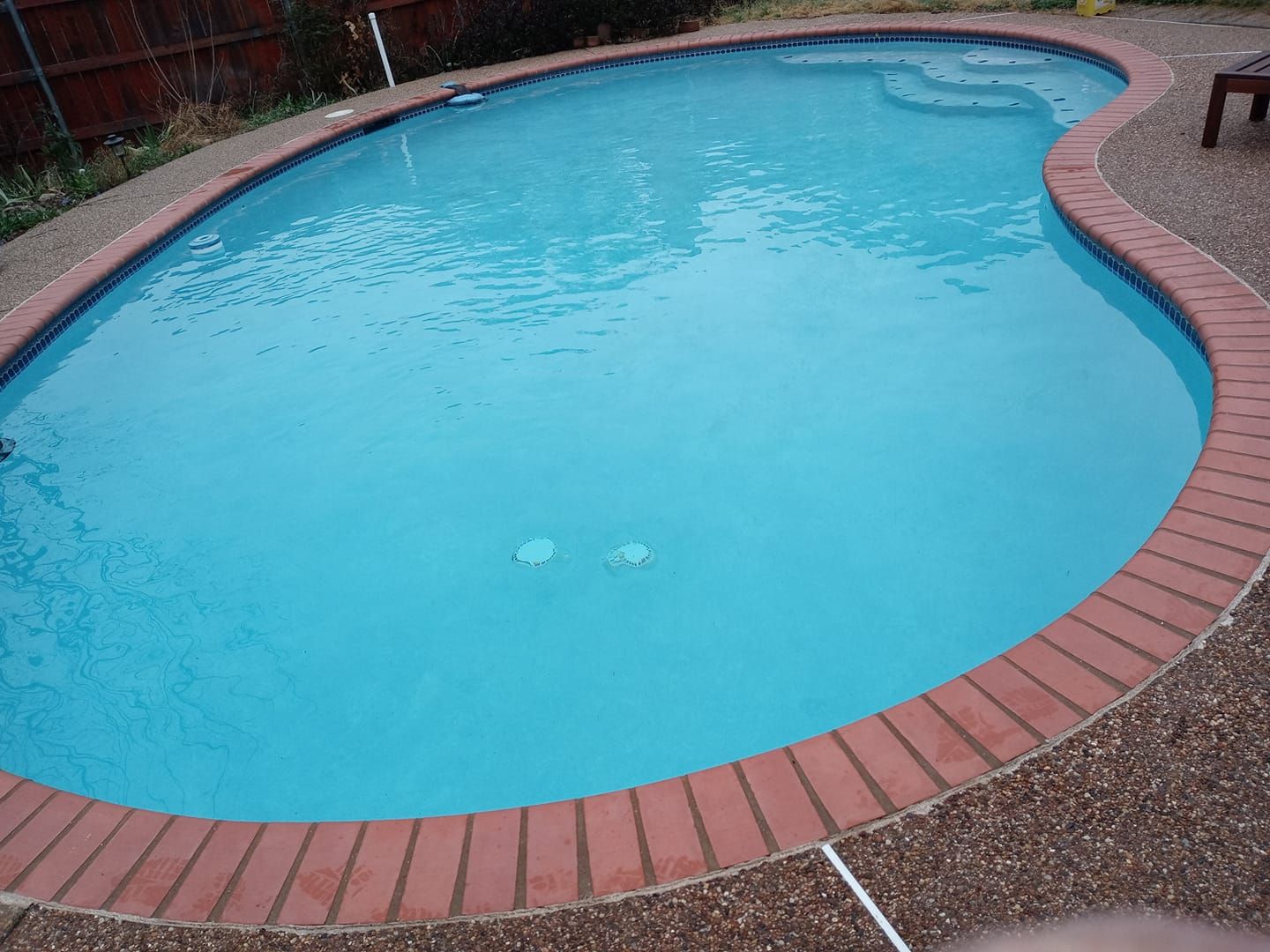 A kidney-shaped swimming pool with blue water and brick edging; a few steps are visible.