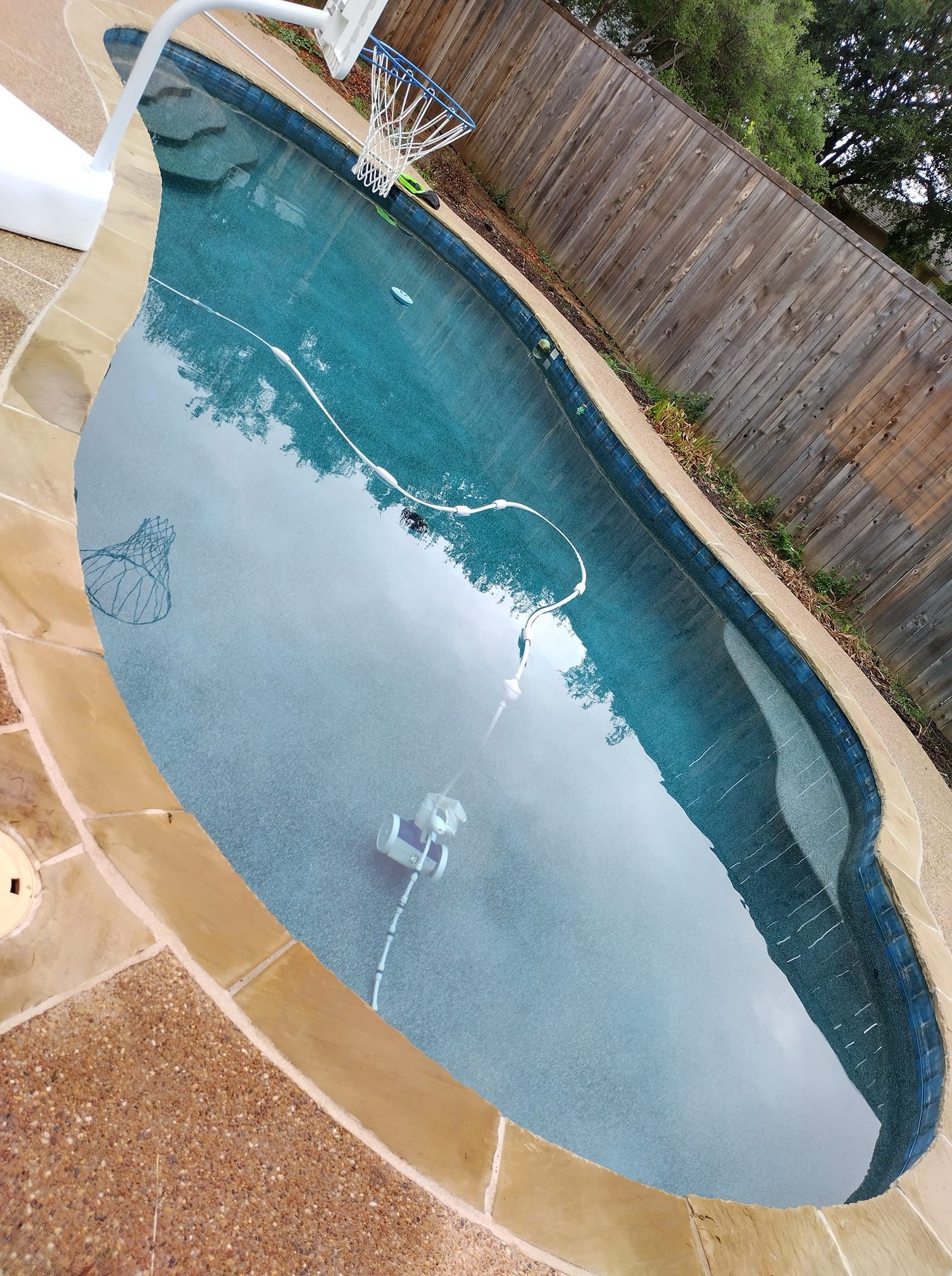 Pool with blue water and stone patio, surrounded by a wooden fence. A pool cleaner is inside.