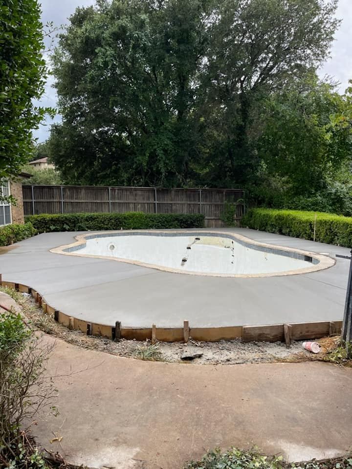 Backyard with a concrete pool under construction; gray concrete, wooden border, green bushes, and a fence.