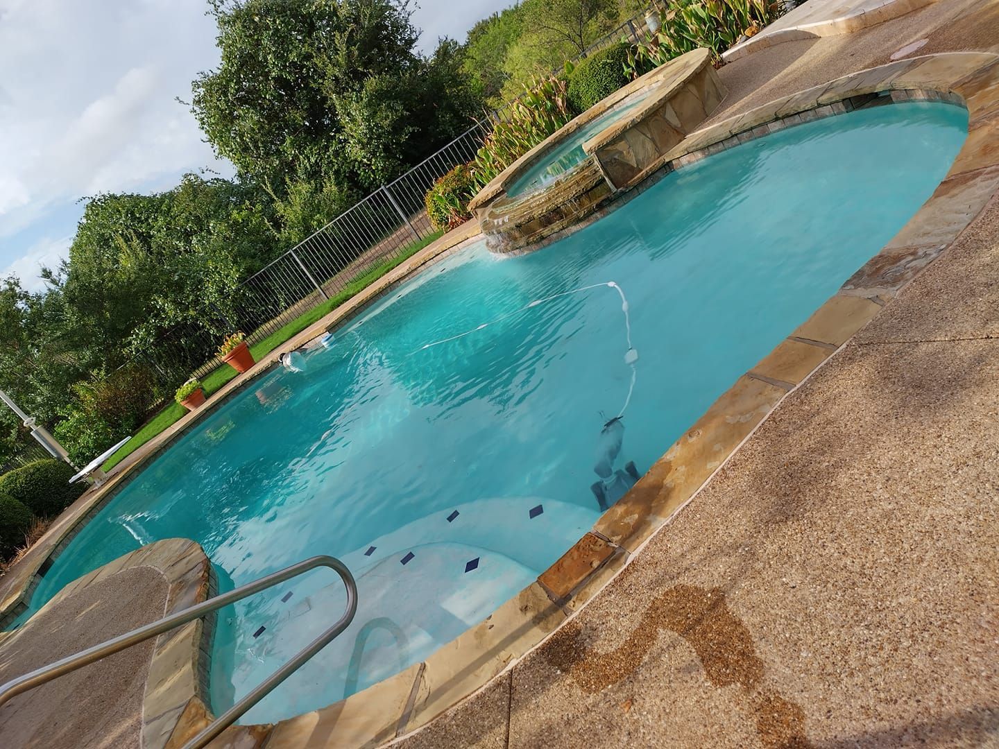Pool with turquoise water, brown concrete deck, and a small waterfall. Green trees in background.