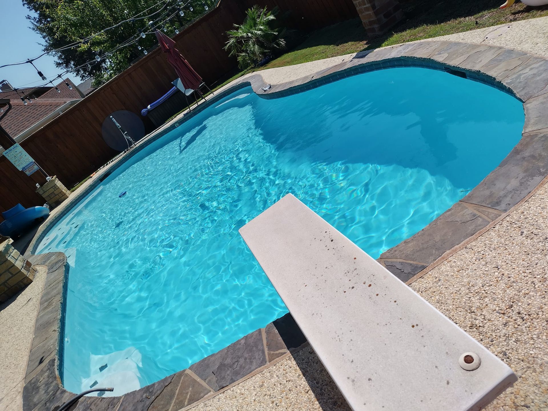 Swimming pool with a diving board, surrounded by stone and a wooden fence. Clear blue water.