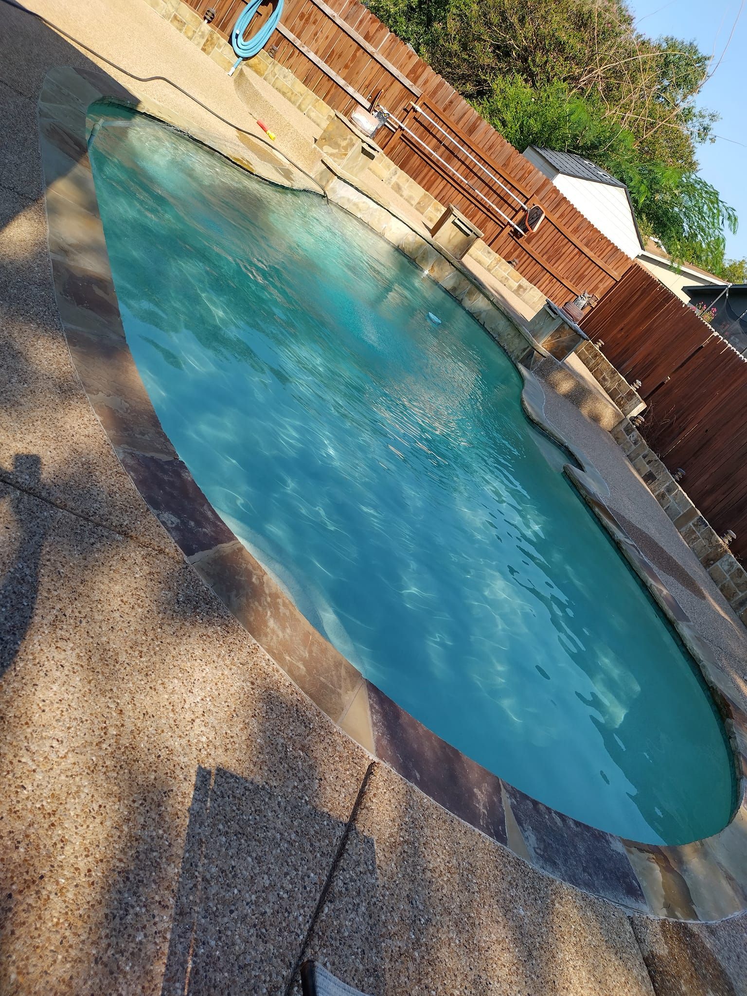 Pool filled with teal water, surrounded by a stone patio and wooden fence under a sunny sky.