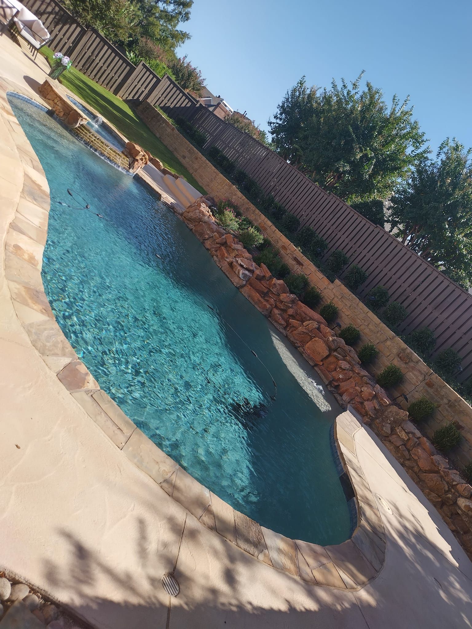 Swimming pool with blue water surrounded by stone and a retaining wall with greenery.