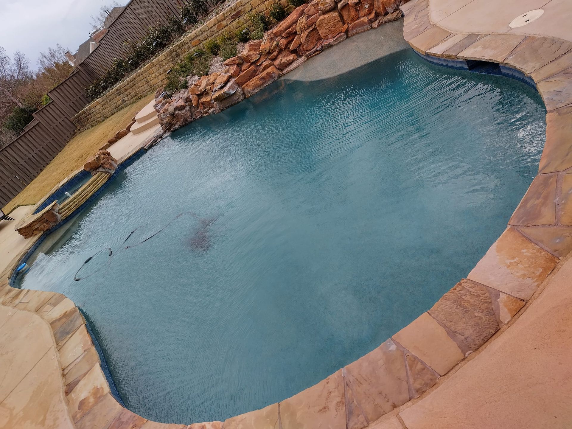 Swimming pool with stone edging and a small rock waterfall feature, in a backyard setting.