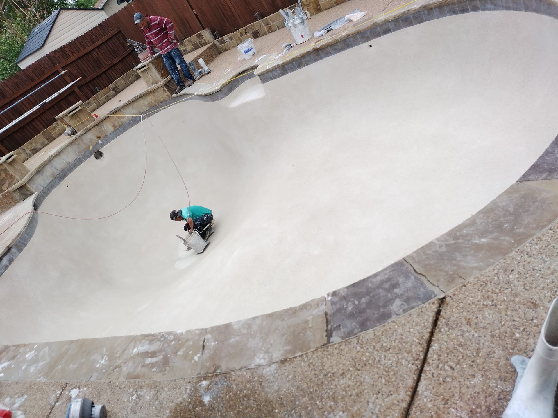 Workers resurfacing a concrete pool. One grinds the base; another stands by the fence.