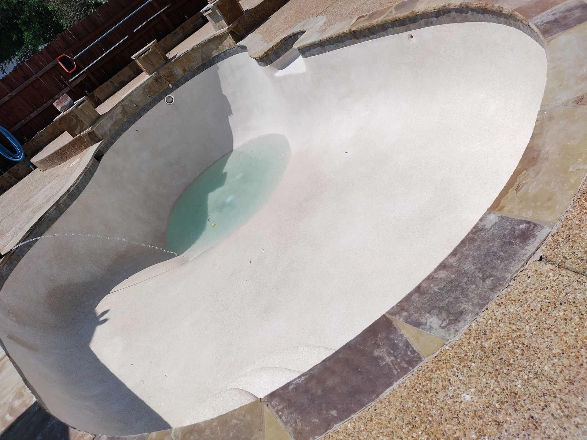 Empty kidney-shaped pool with light-colored plaster, surrounded by stone and concrete patio.