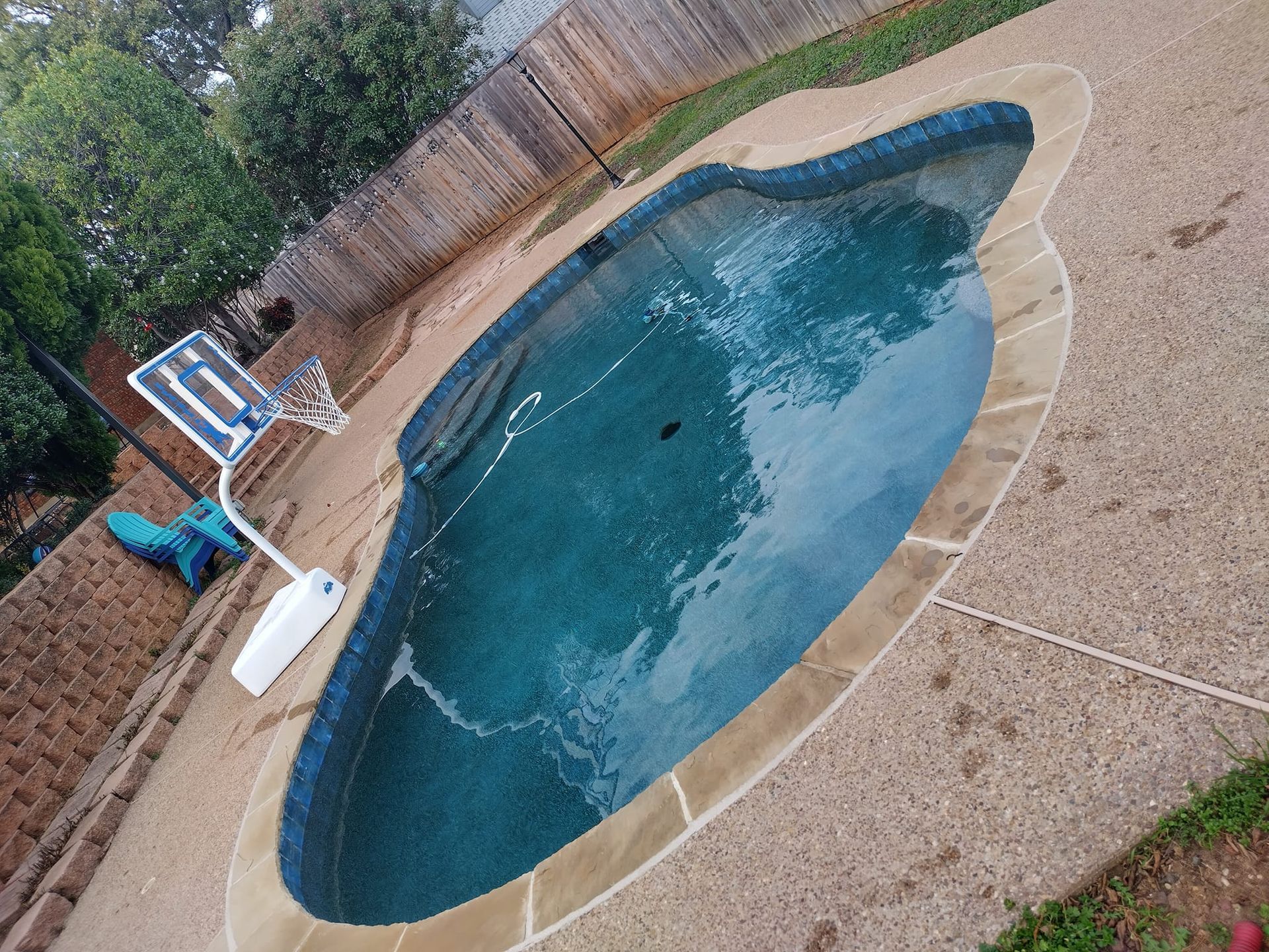 Swimming pool with teal water, concrete deck, basketball hoop, wooden fence.