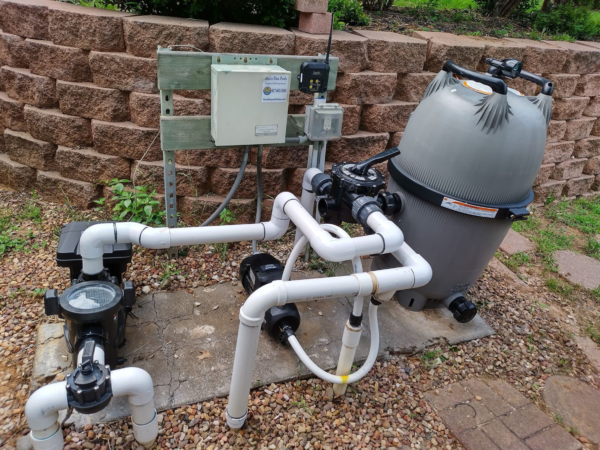 Pool equipment setup: pump, filter, plumbing, and control box against a brick retaining wall.