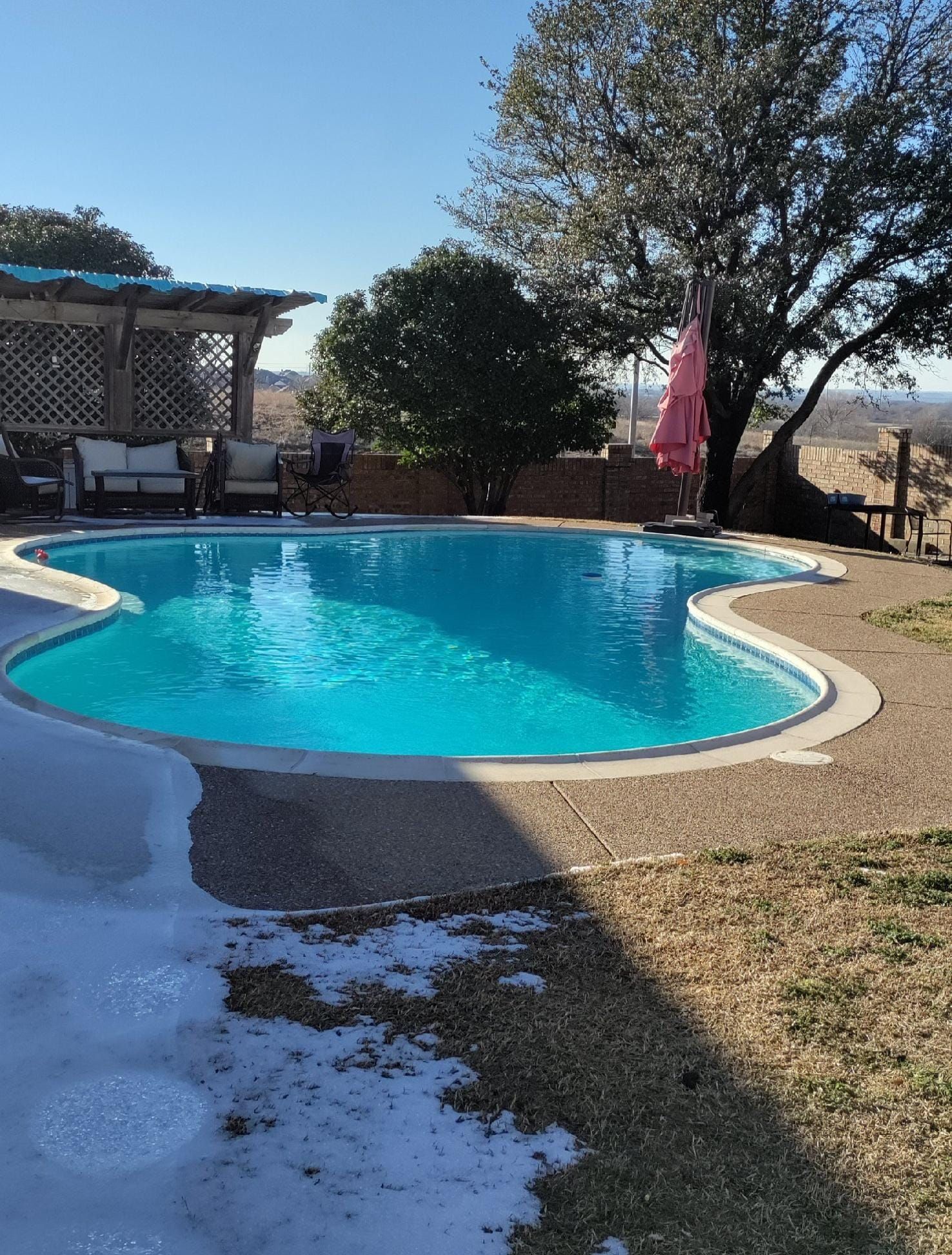 A curvy turquoise pool sits next to snow and grass. A pergola and umbrella are nearby.