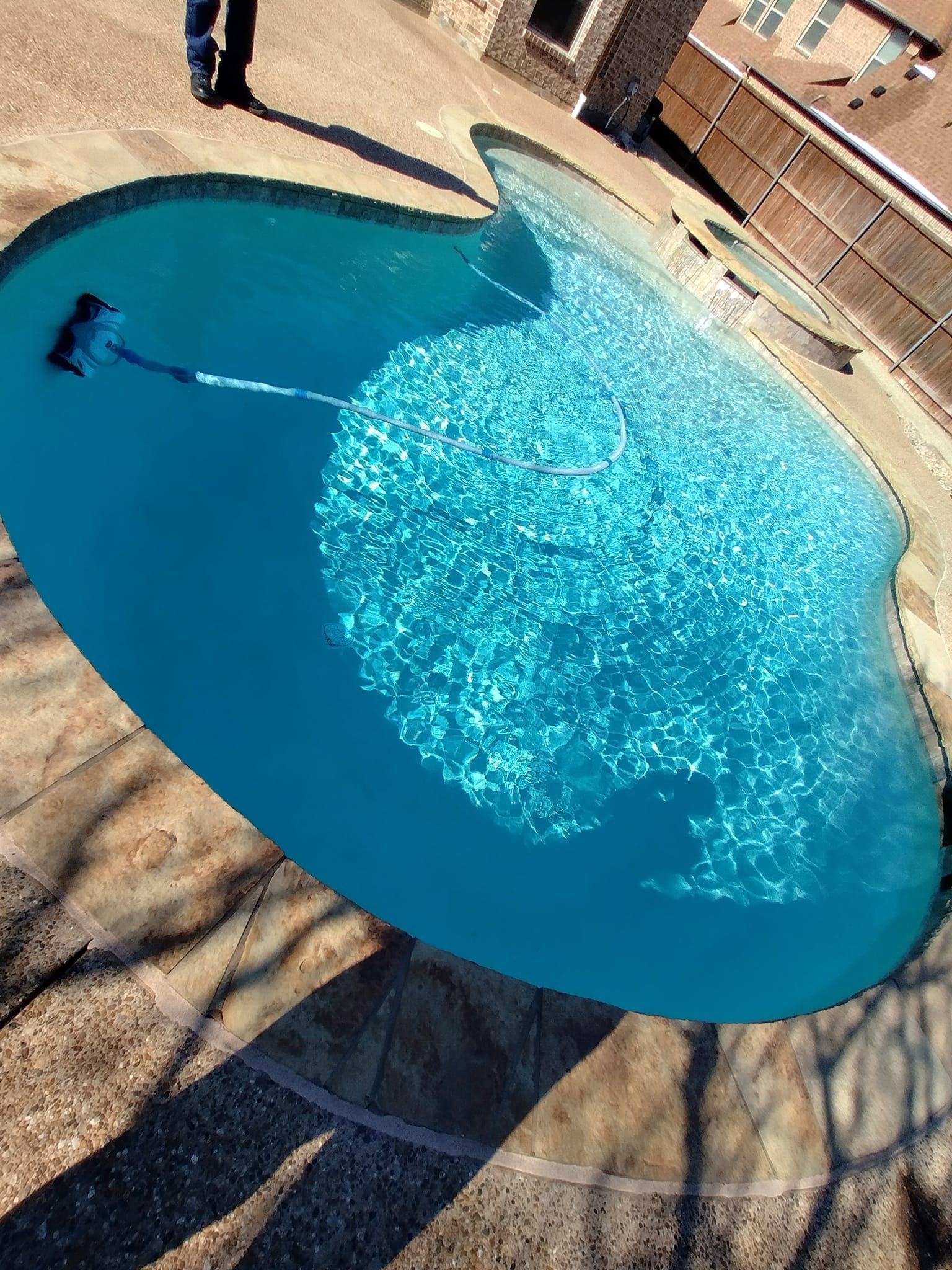 Pool being cleaned with a blue water surface, person's legs visible near edge, stone patio and wooden fence.