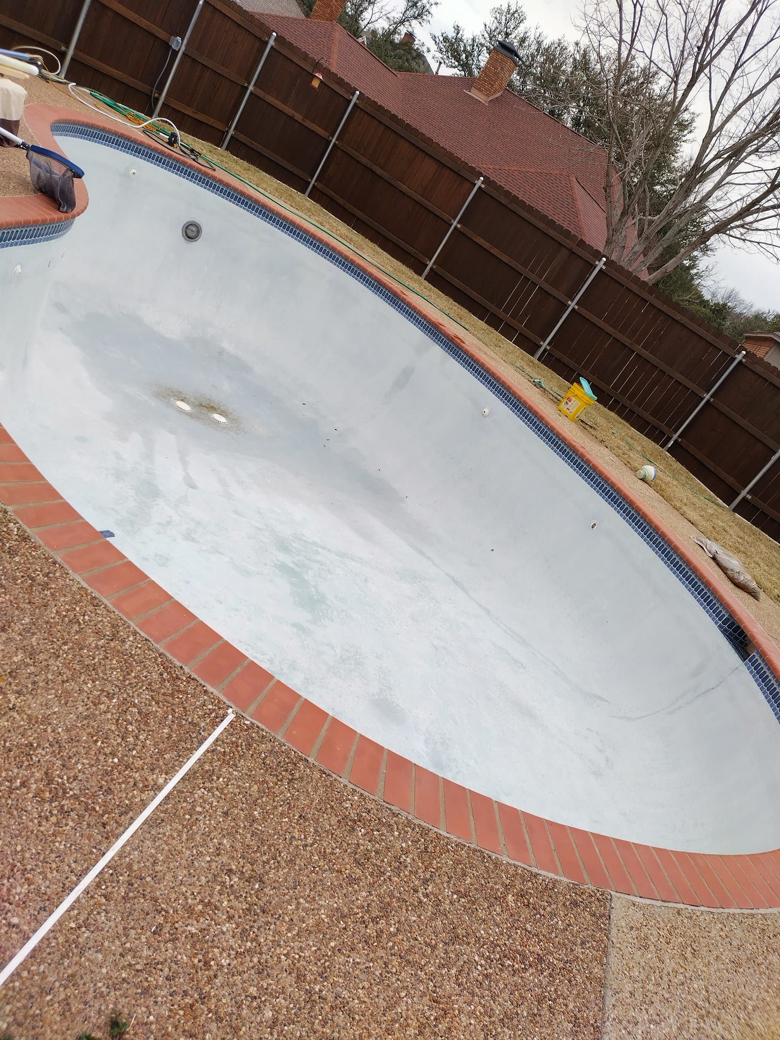 Empty pool with brown brick border, tan speckled concrete, and a brown fence in the background.