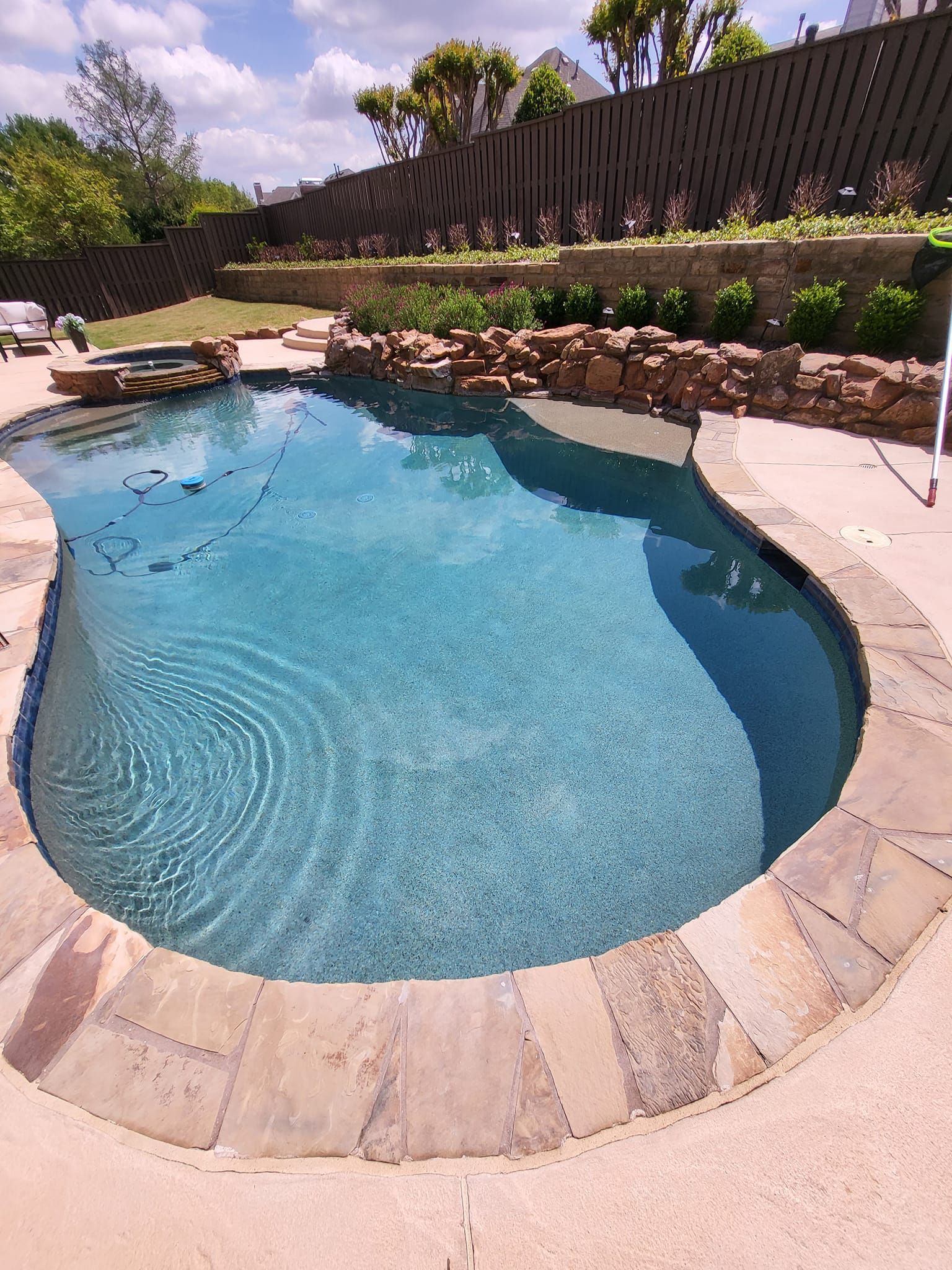 A blue swimming pool with stone coping, a small hot tub, and a pool cleaner.