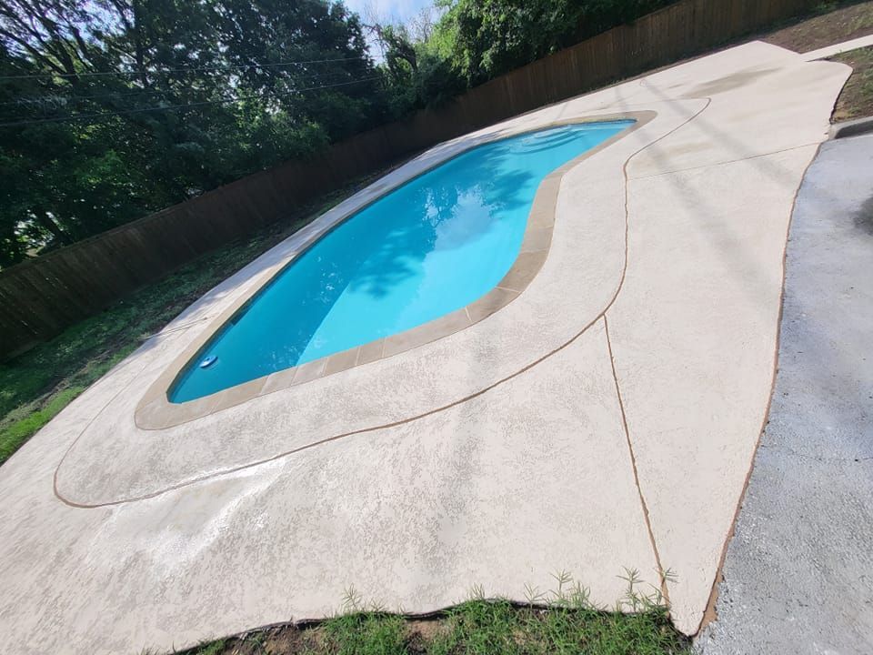 A kidney-shaped swimming pool with blue water surrounded by concrete, next to grass and a wooden fence.