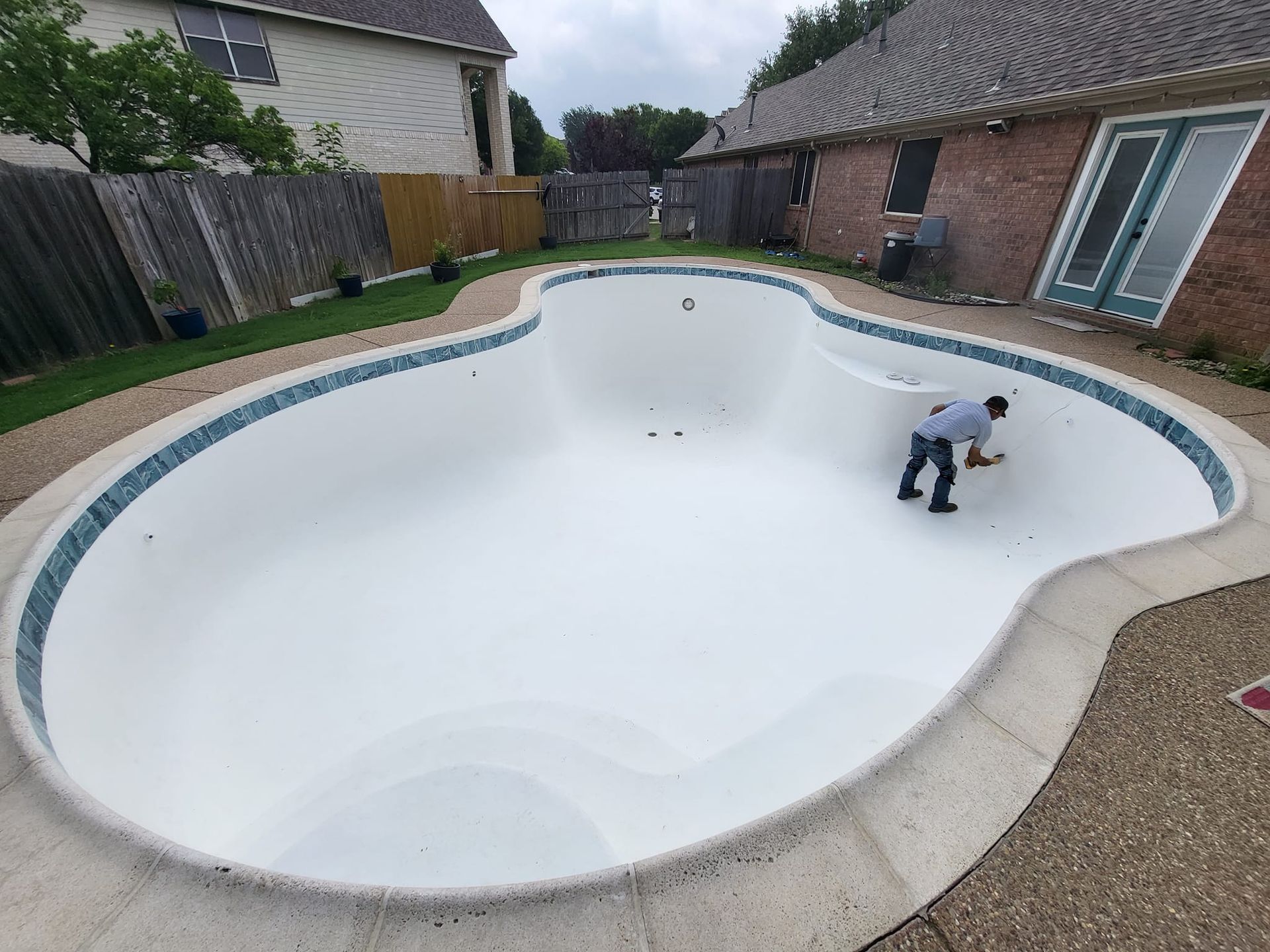Man cleaning an empty, kidney-shaped swimming pool. The pool has a blue-tiled border and is surrounded by a patio and yard.