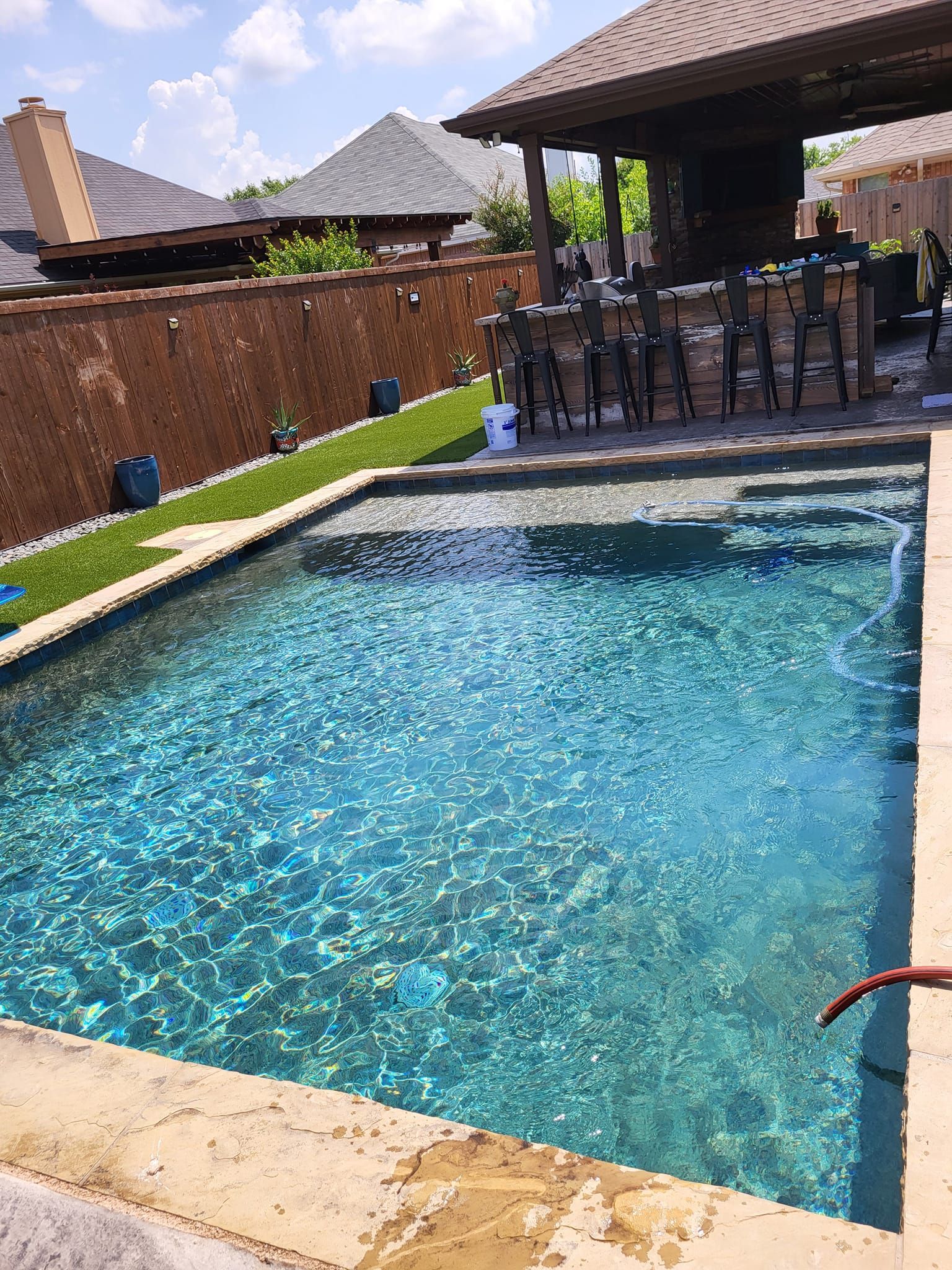 Pool with blue water and stone patio, adjacent to a covered bar area and brick wall.