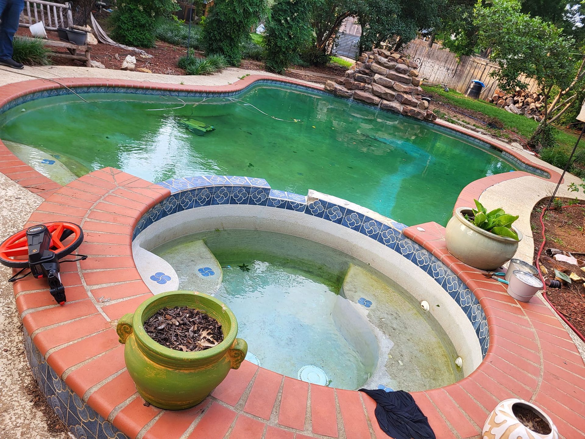 Green algae-filled pool and spa surrounded by brick and stone, small potted plants visible.