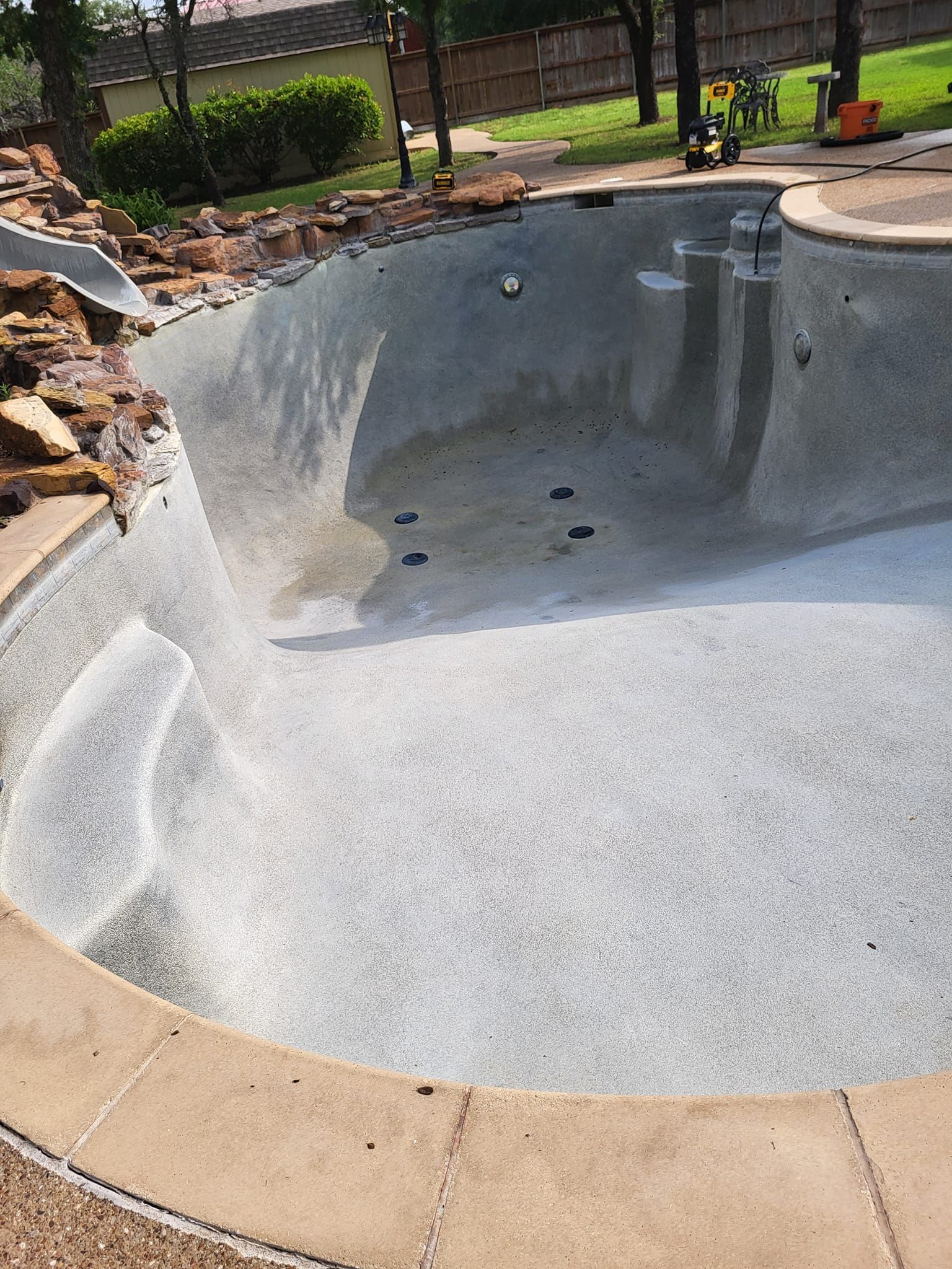 Empty swimming pool with gray interior, waterfall feature, and tan concrete border, outdoors.