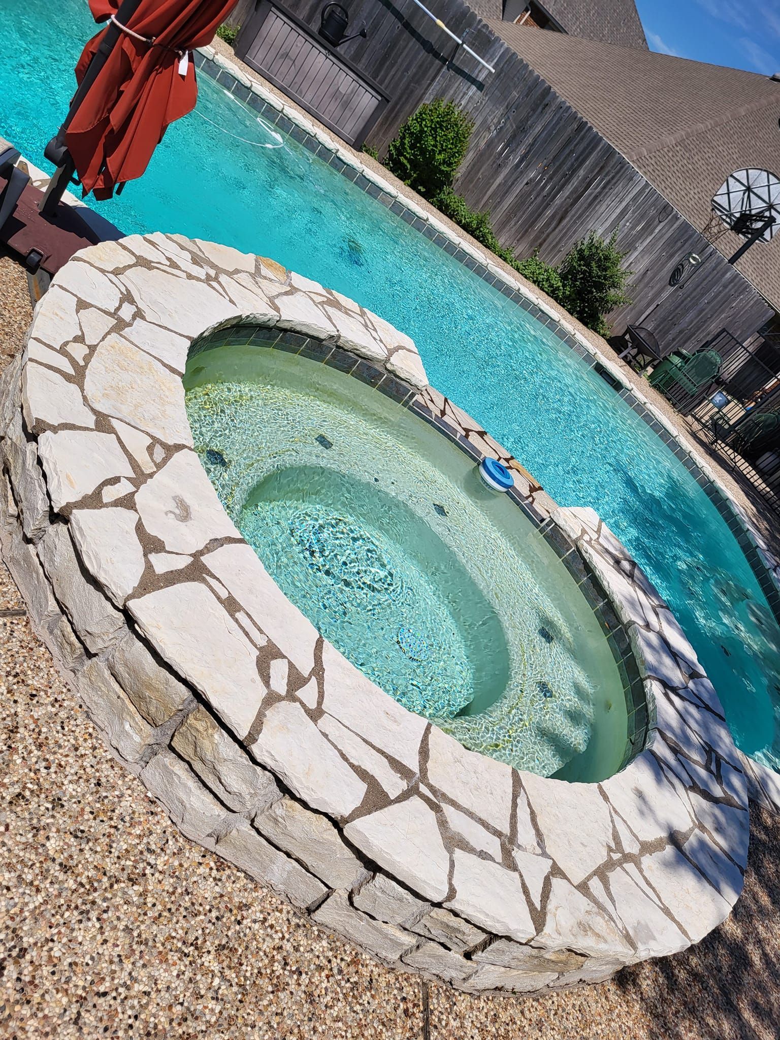 Swimming pool with built-in spa. Stone surround, clear water. Brown patio. Red umbrella.