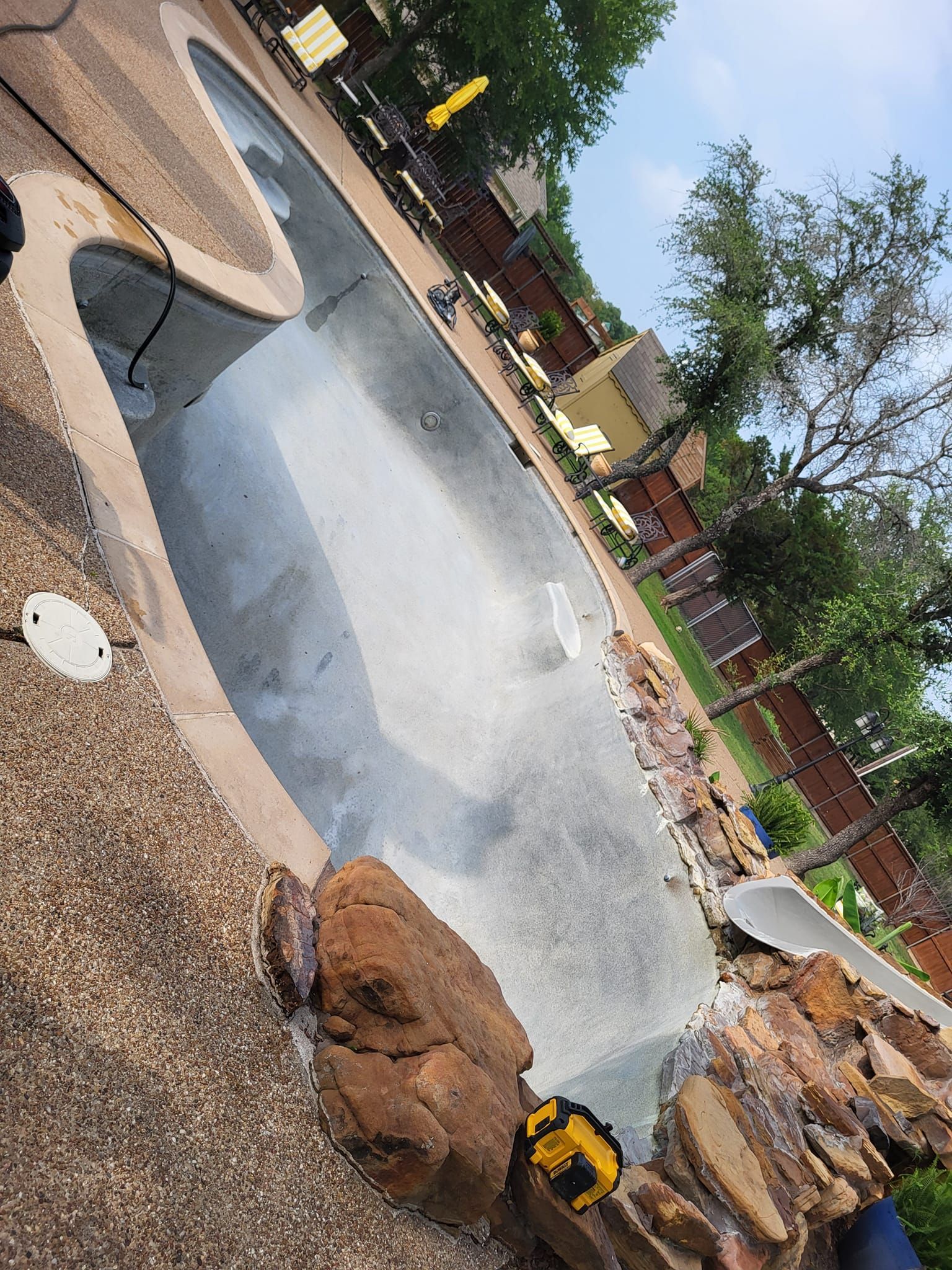 Pool with dark gray surface, surrounded by textured stone and landscaping under a blue sky.