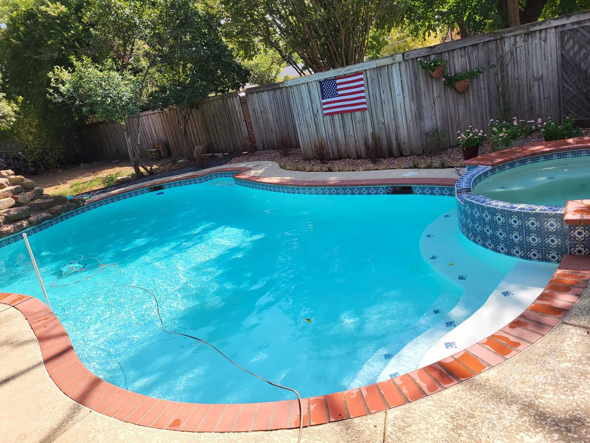 Pool with turquoise water, brick border, and hot tub. American flag on wooden fence in background.