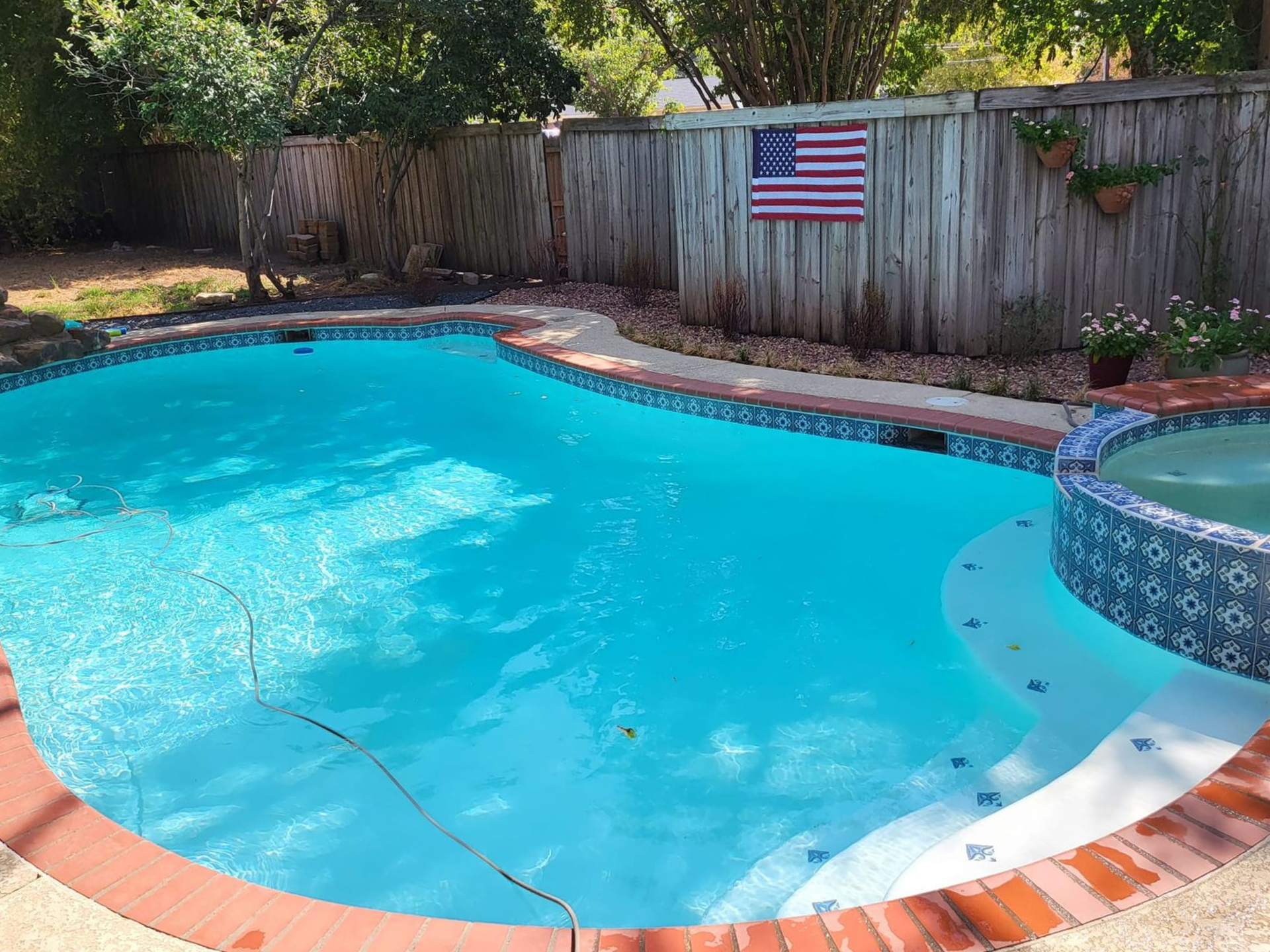 Swimming pool with stone coping and jacuzzi, next to a rock waterfall feature; green surroundings.