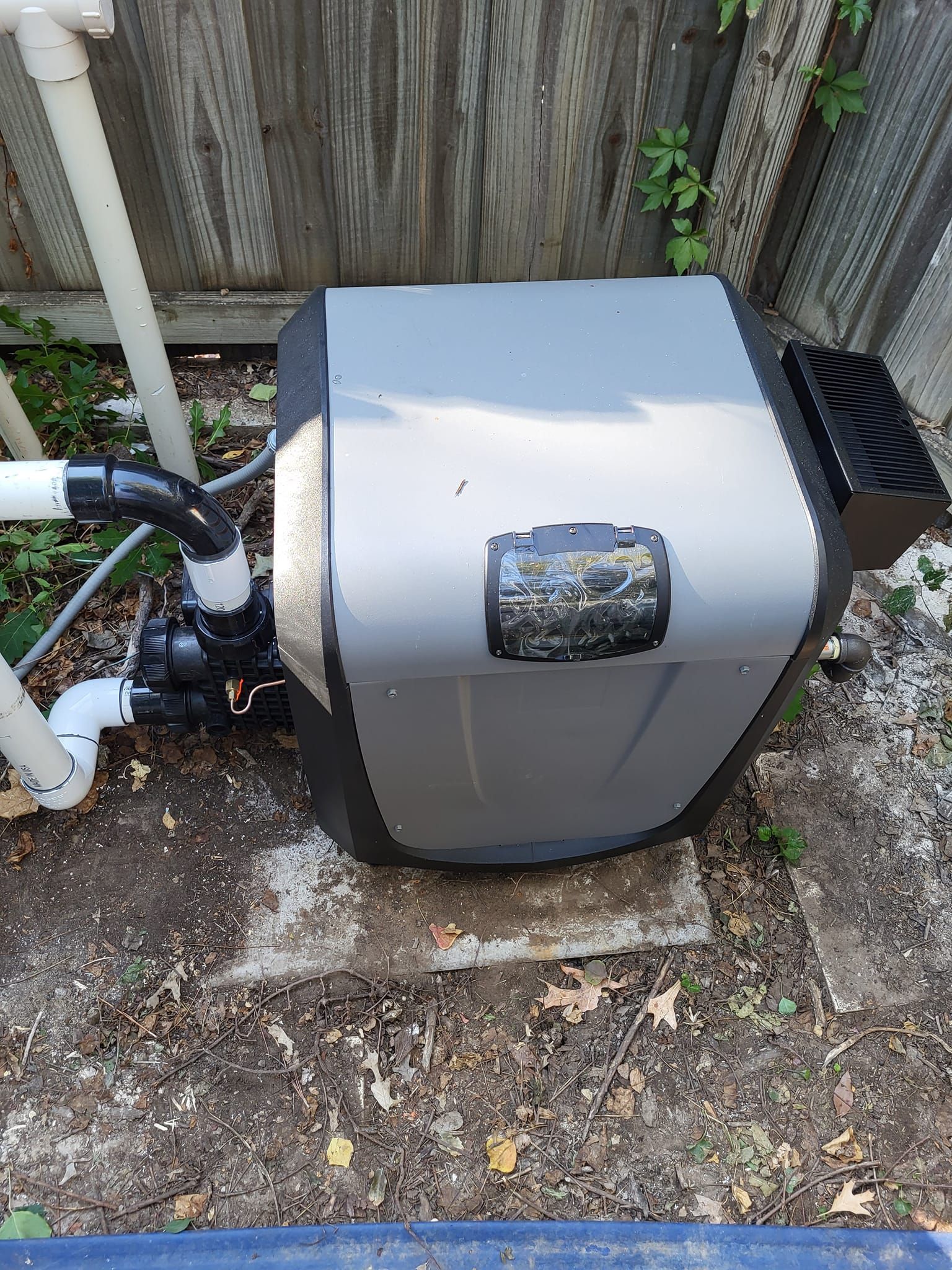 A pool heater and pump, gray and black, next to pipes, dirt, and a wooden fence.
