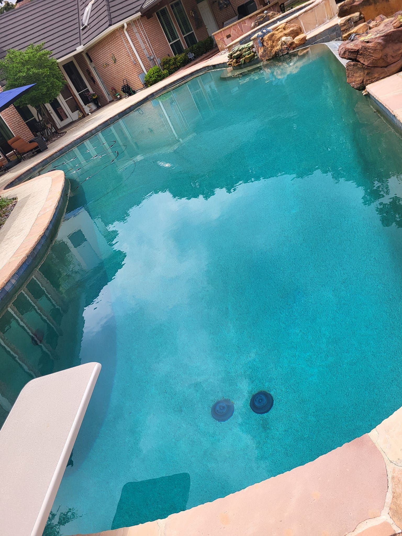Swimming pool with a diving board, surrounded by stone patio and building. Turquoise water.