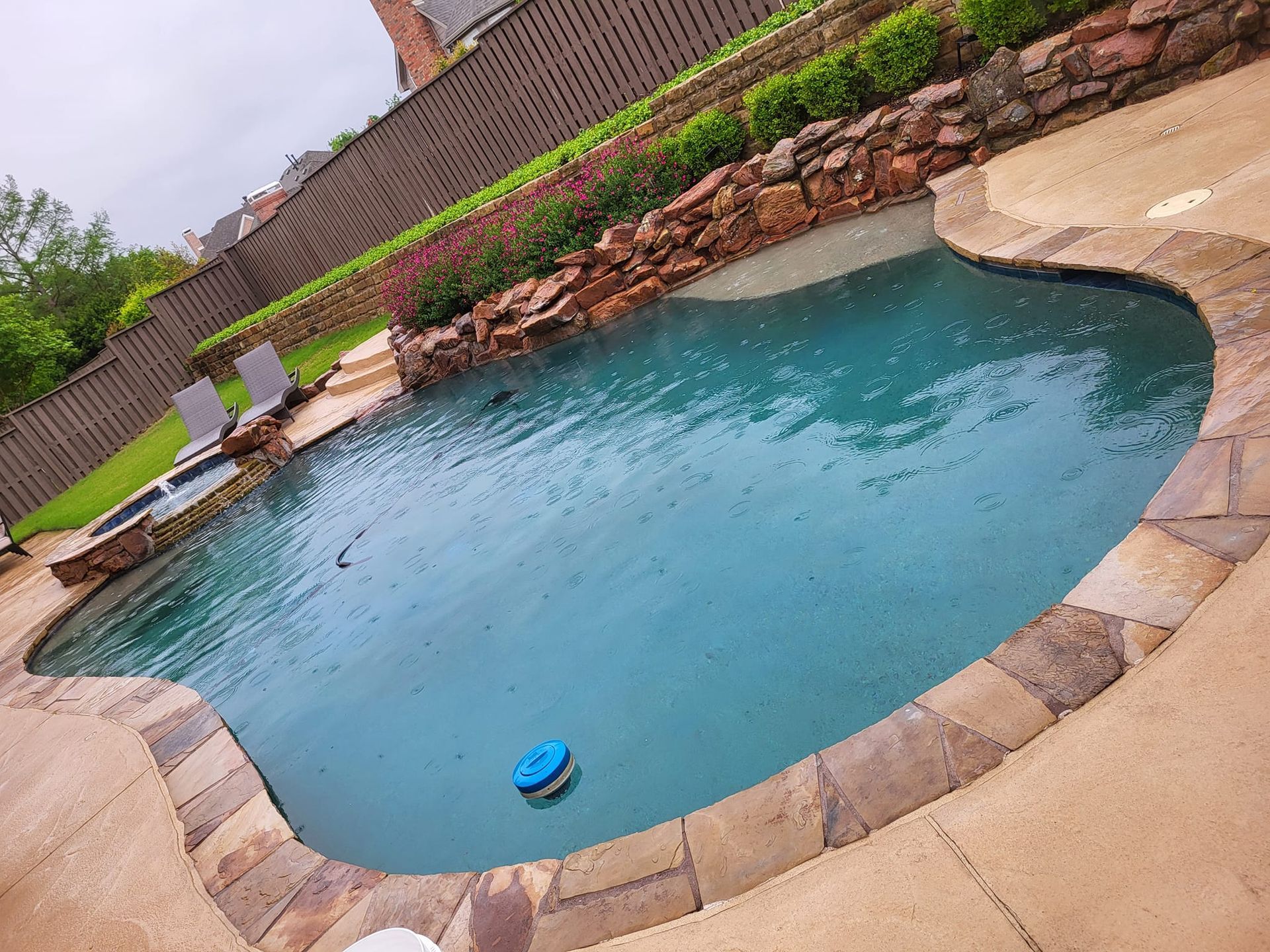 Swimming pool with stone edging. Rocks and plants border one side. Gray sky.