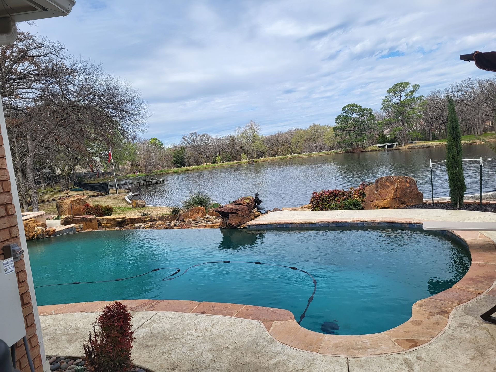 Swimming pool overlooking a lake, with rocky landscaping and blue water under a cloudy sky.
