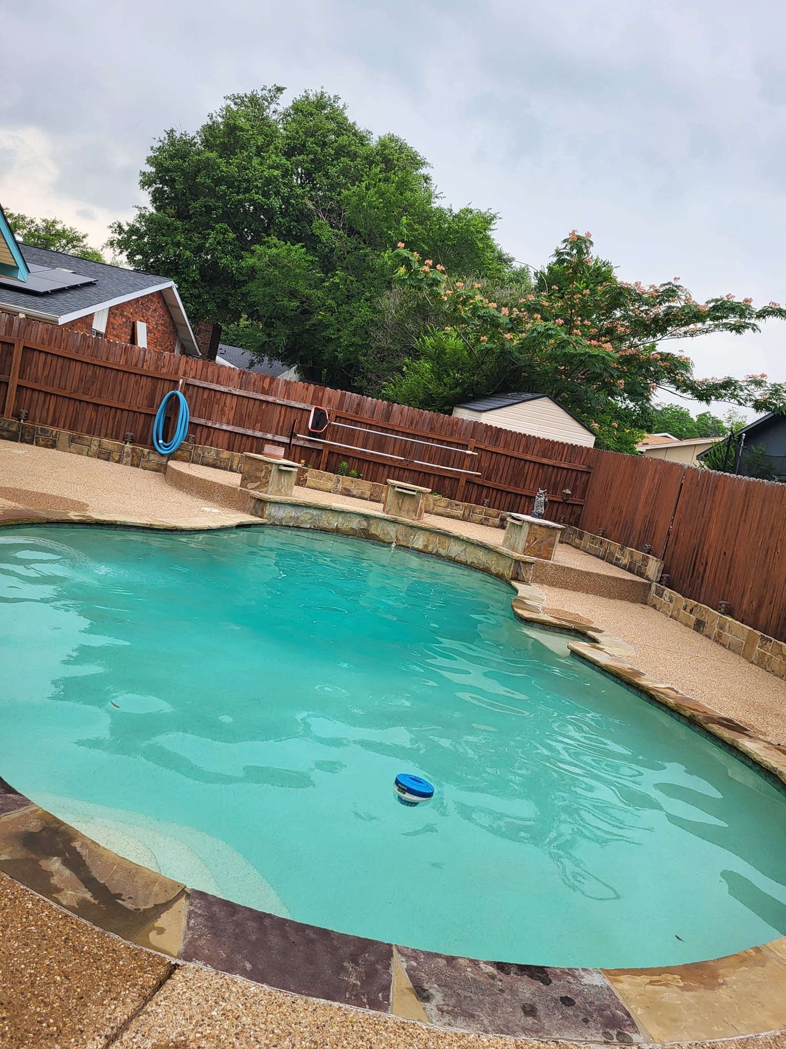 Swimming pool with turquoise water, surrounded by a brown stone deck and a wooden fence. Overcast sky.