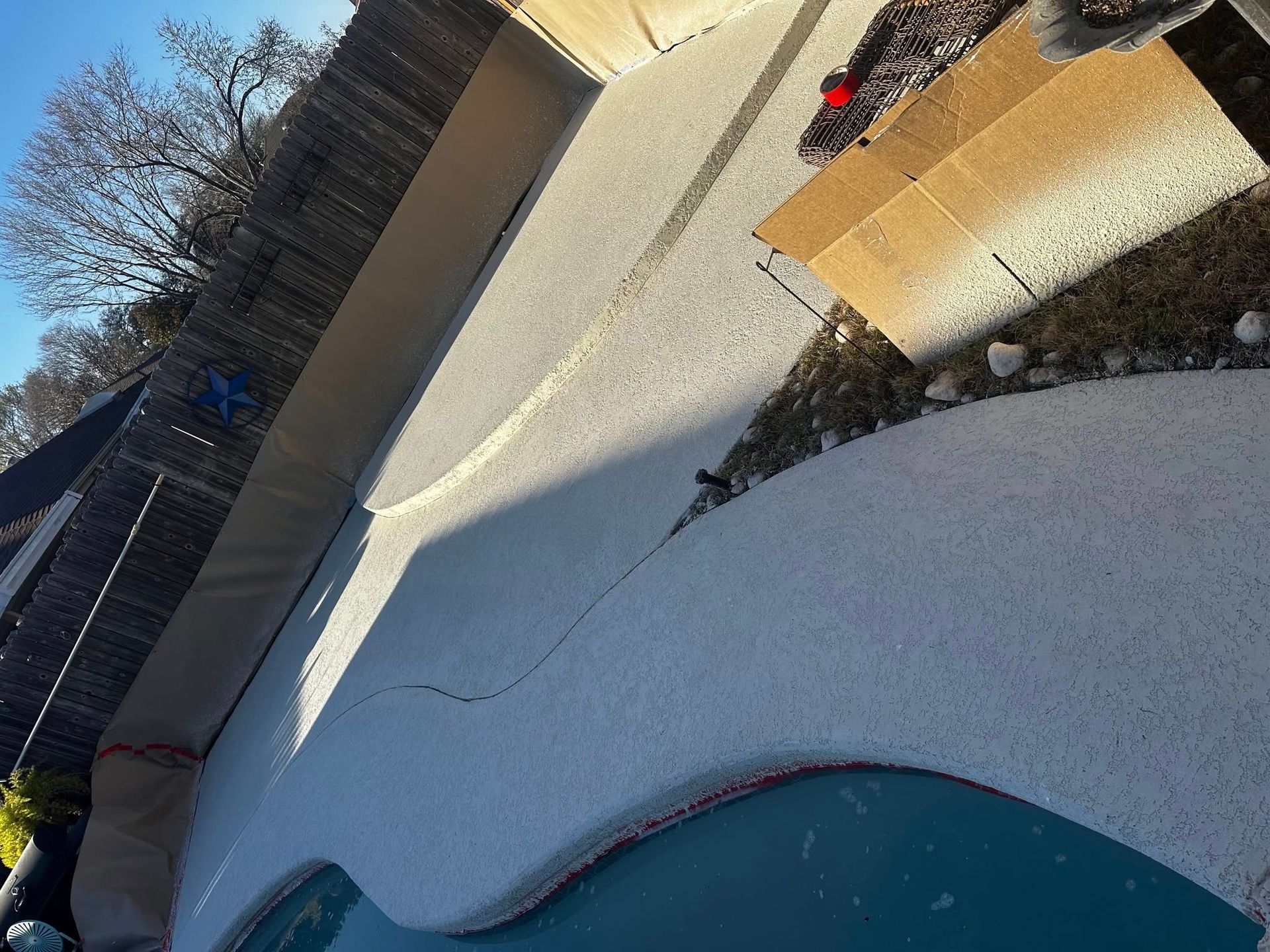 Pool and patio area with light-colored concrete. The pool water is blue. A brick fence is in the background.
