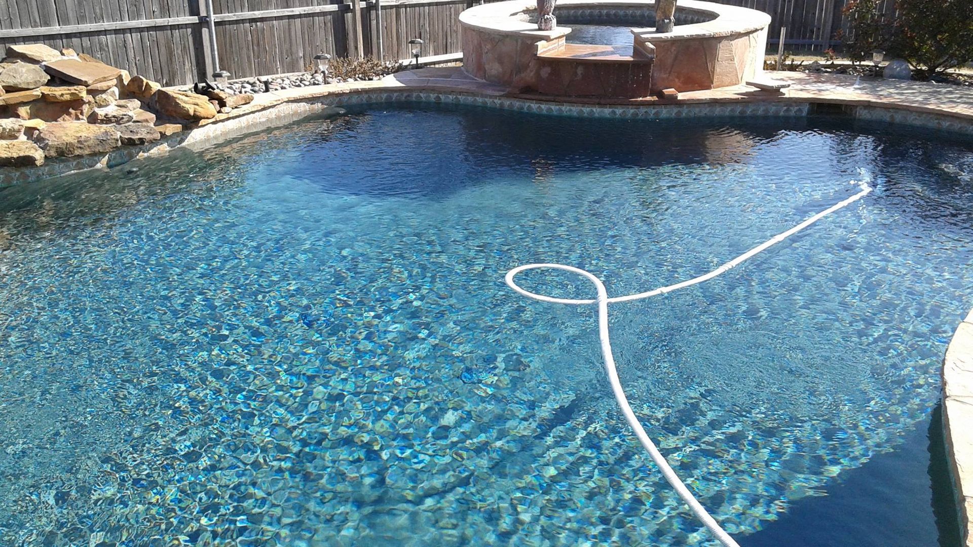 Swimming pool with clear blue water and attached hot tub. A pool hose floats in the water.