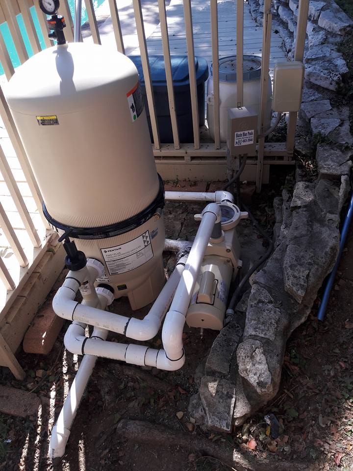 Pool filter system with a beige tank and white pipes, located near a deck and rocks.