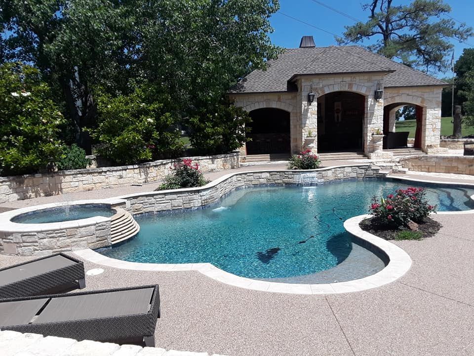 Swimming pool with stone features, a spa, and a small building under a blue sky.