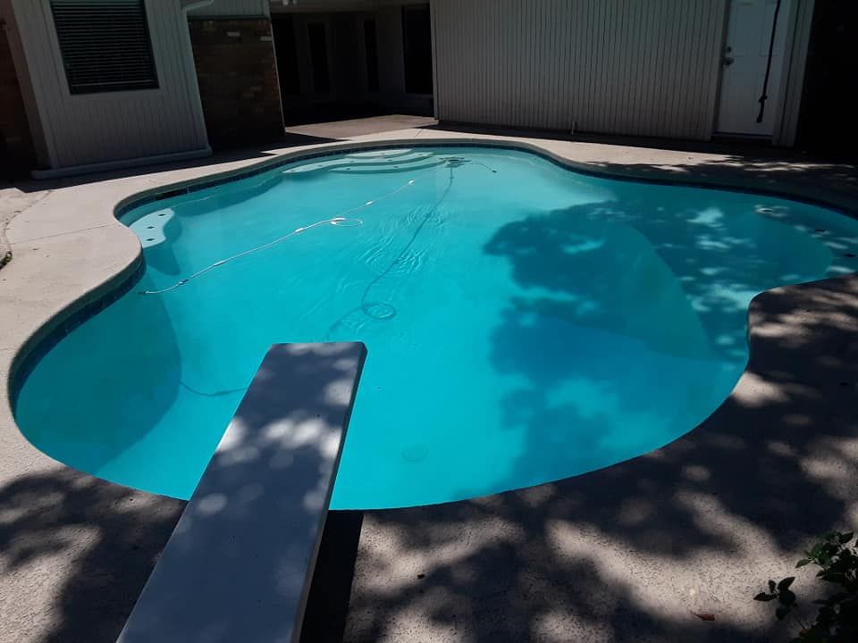 Pool with diving board and light blue water, surrounded by concrete and trees.