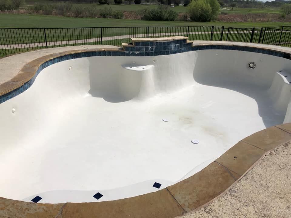 Empty kidney-shaped swimming pool with beige concrete border, blue tile trim, and black fence in background.