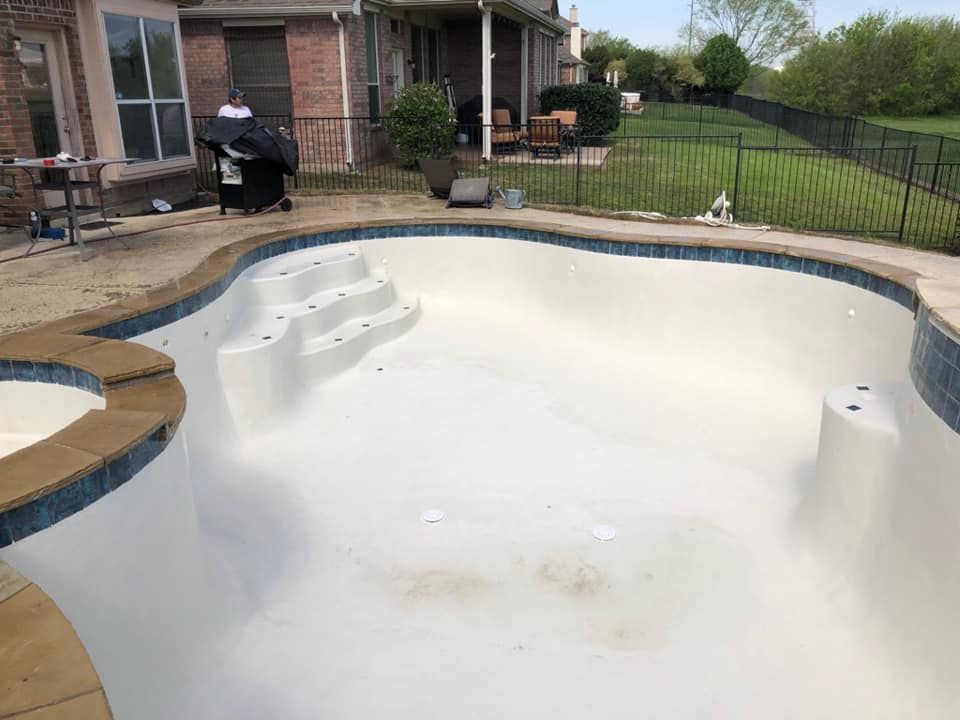 Empty, white-tiled swimming pool with blue tile border, steps, and surrounding patio area. Houses in the background.
