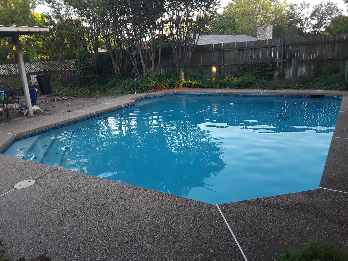 Pool with blue water and concrete border surrounded by a fence and trees.