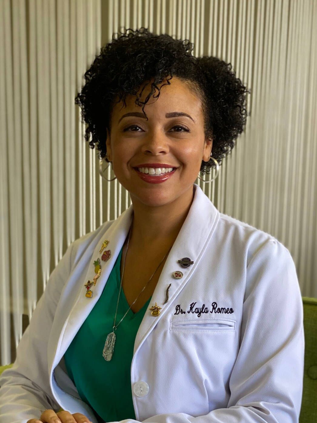A woman wearing a white lab coat and a green shirt is smiling.