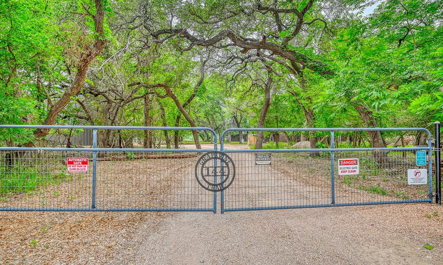 A large metal gate is surrounded by trees on a gravel road.