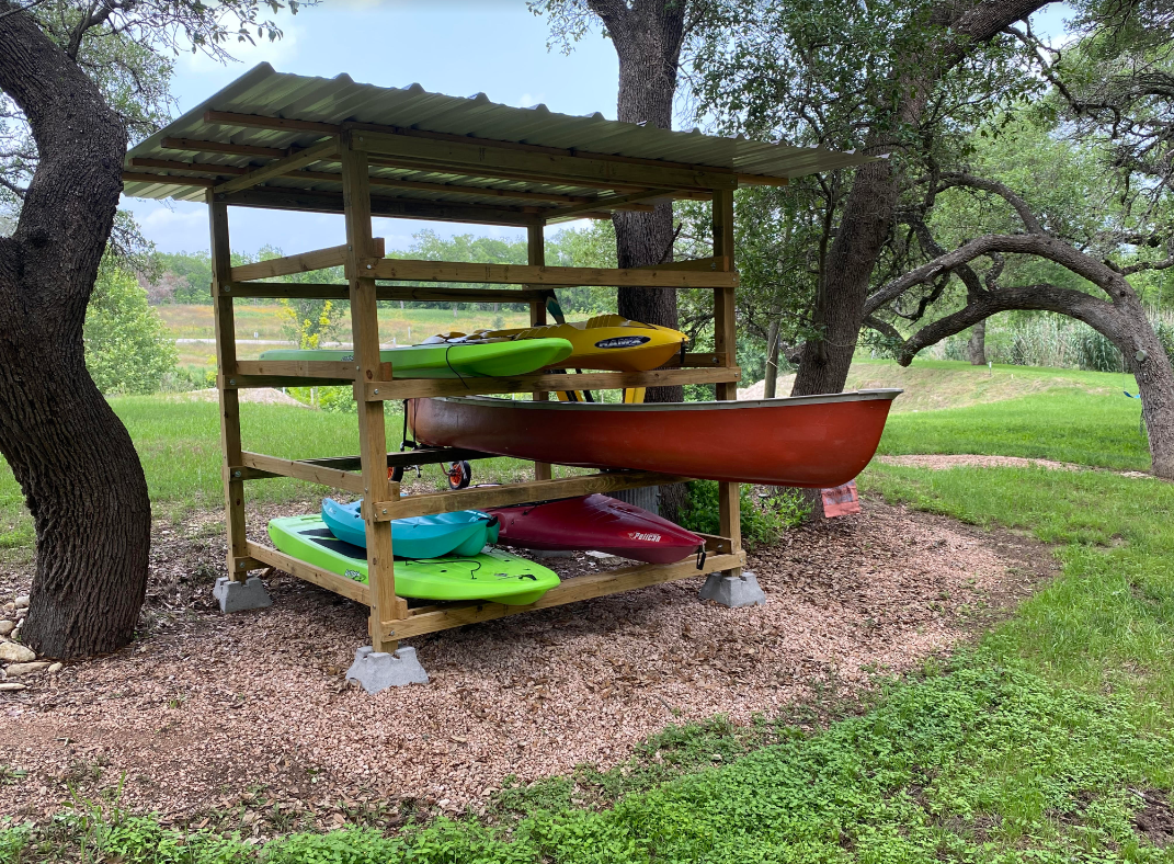 A wooden shelter with kayaks inside of it is sitting under a tree.