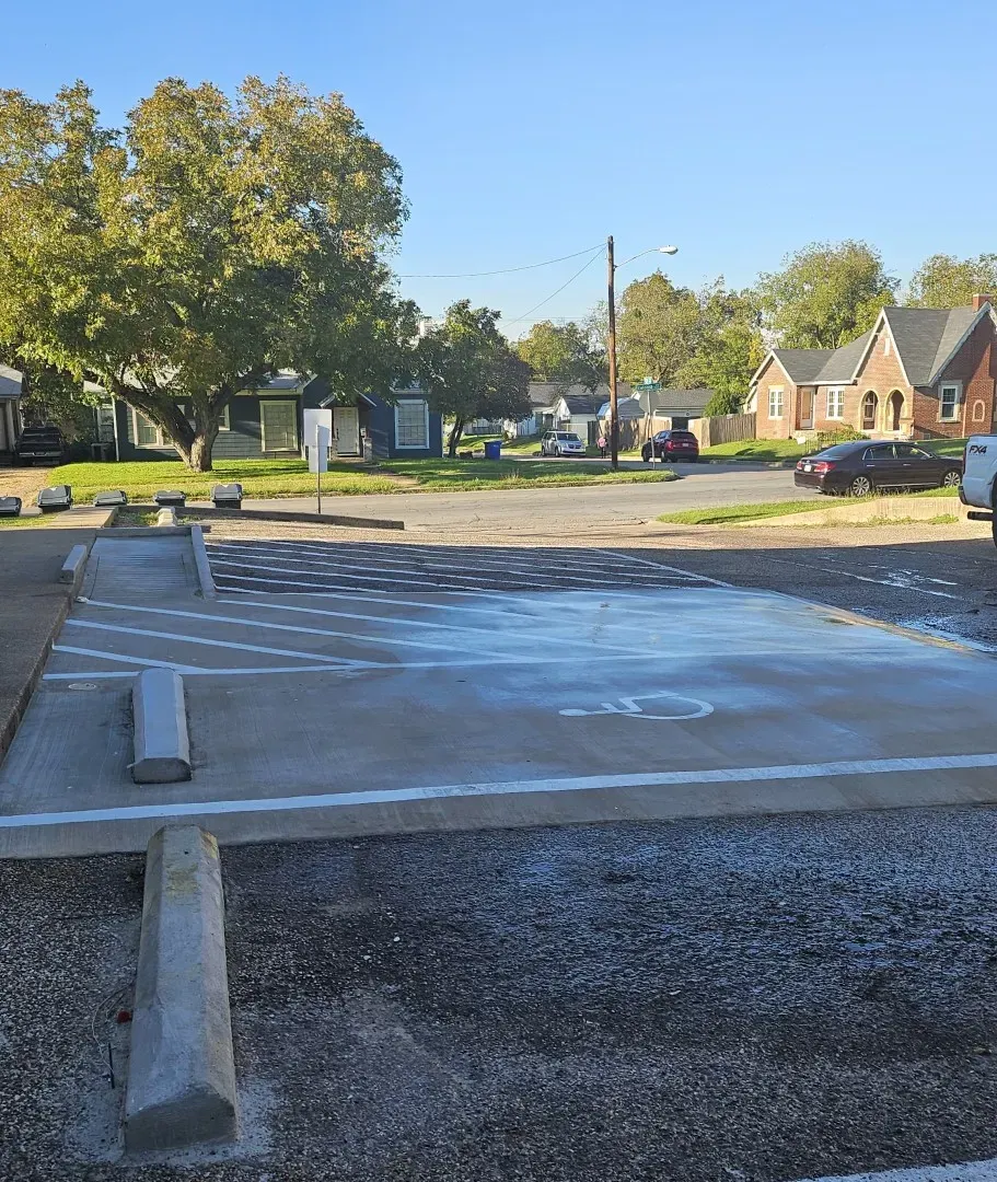 Empty concrete foundation slab at a residential construction site, with gravel, trees, and houses in the background