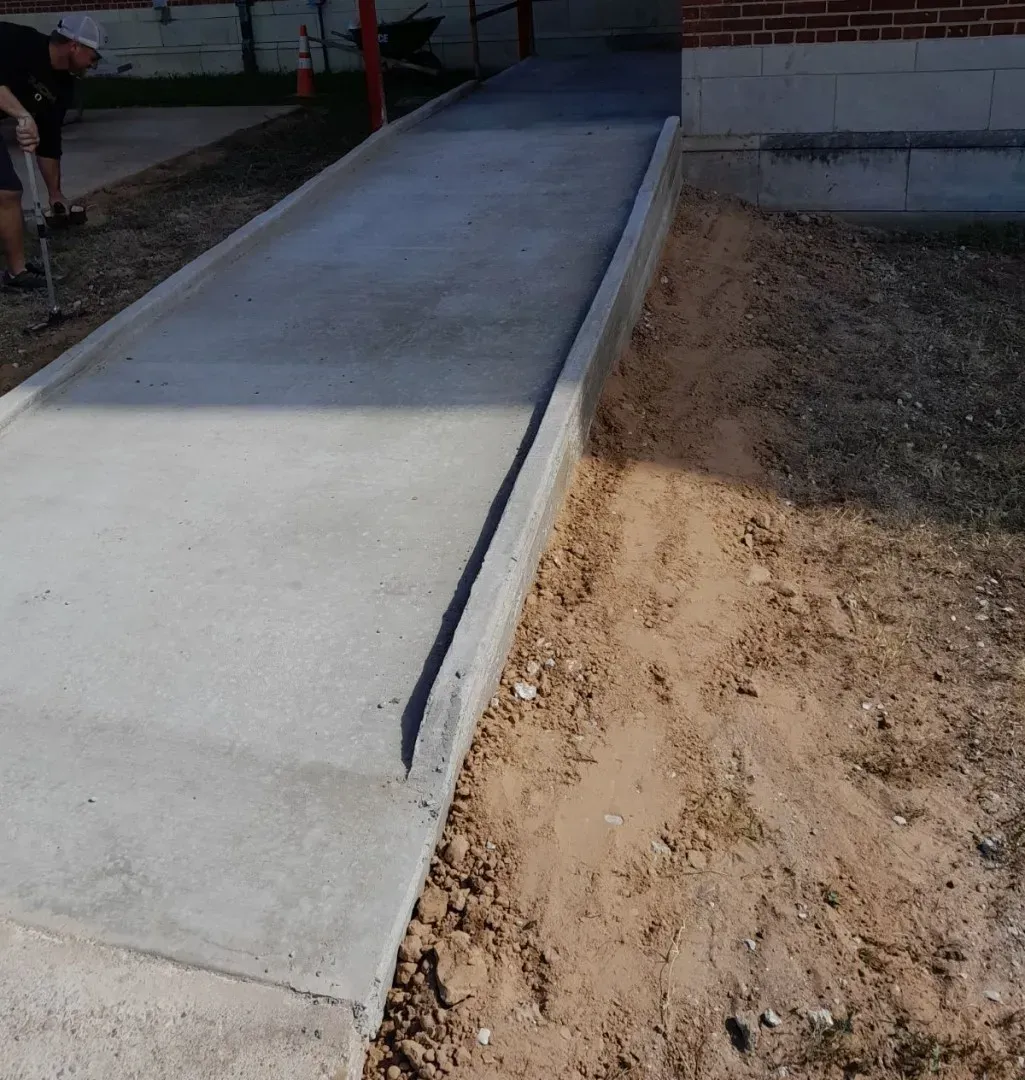 Concrete wheelchair ramp beside a brick building, with unfinished dirt ground along one side.
