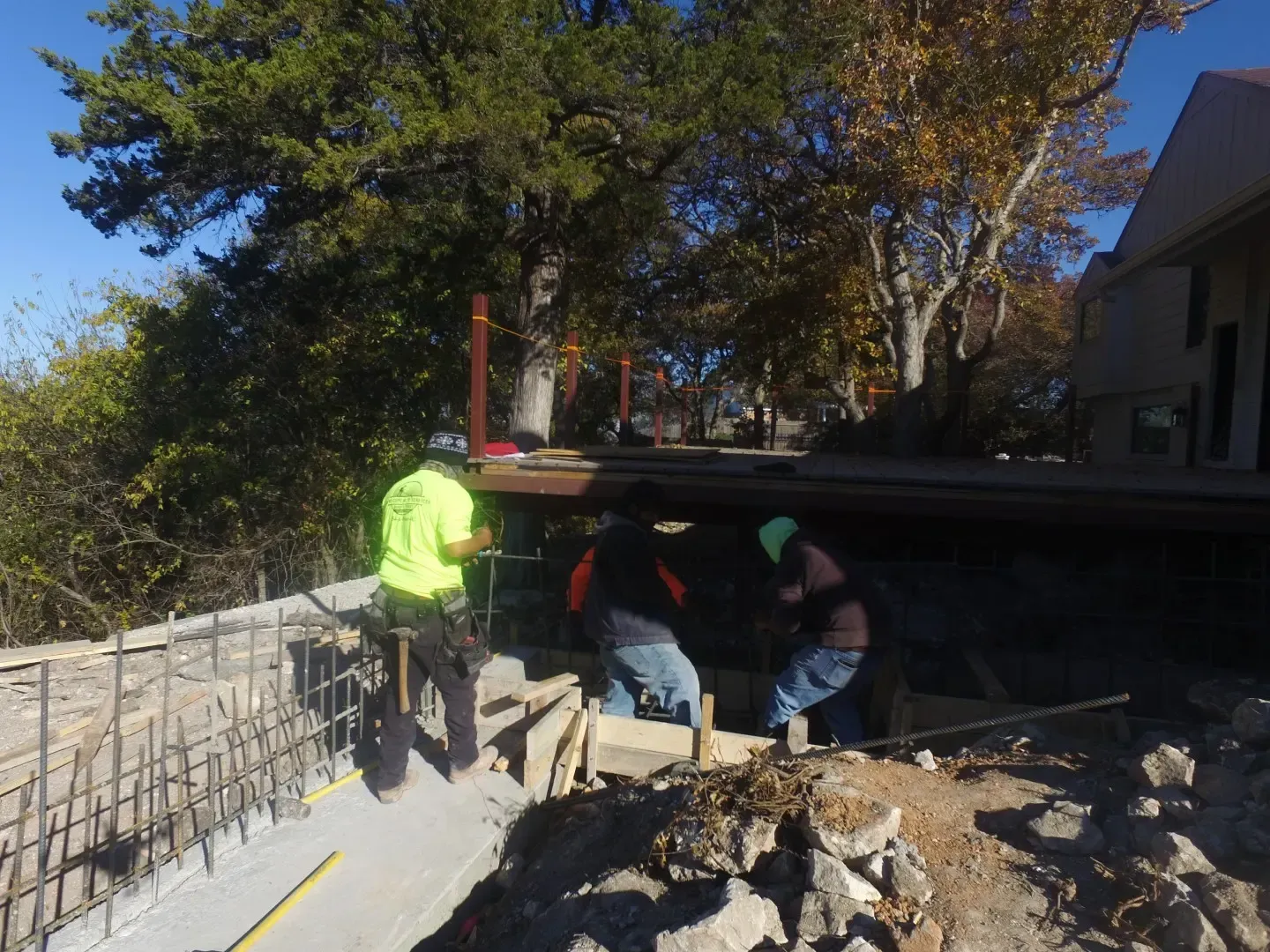 Workers in safety vests repair a collapsed sidewalk near a house under a bridge.
