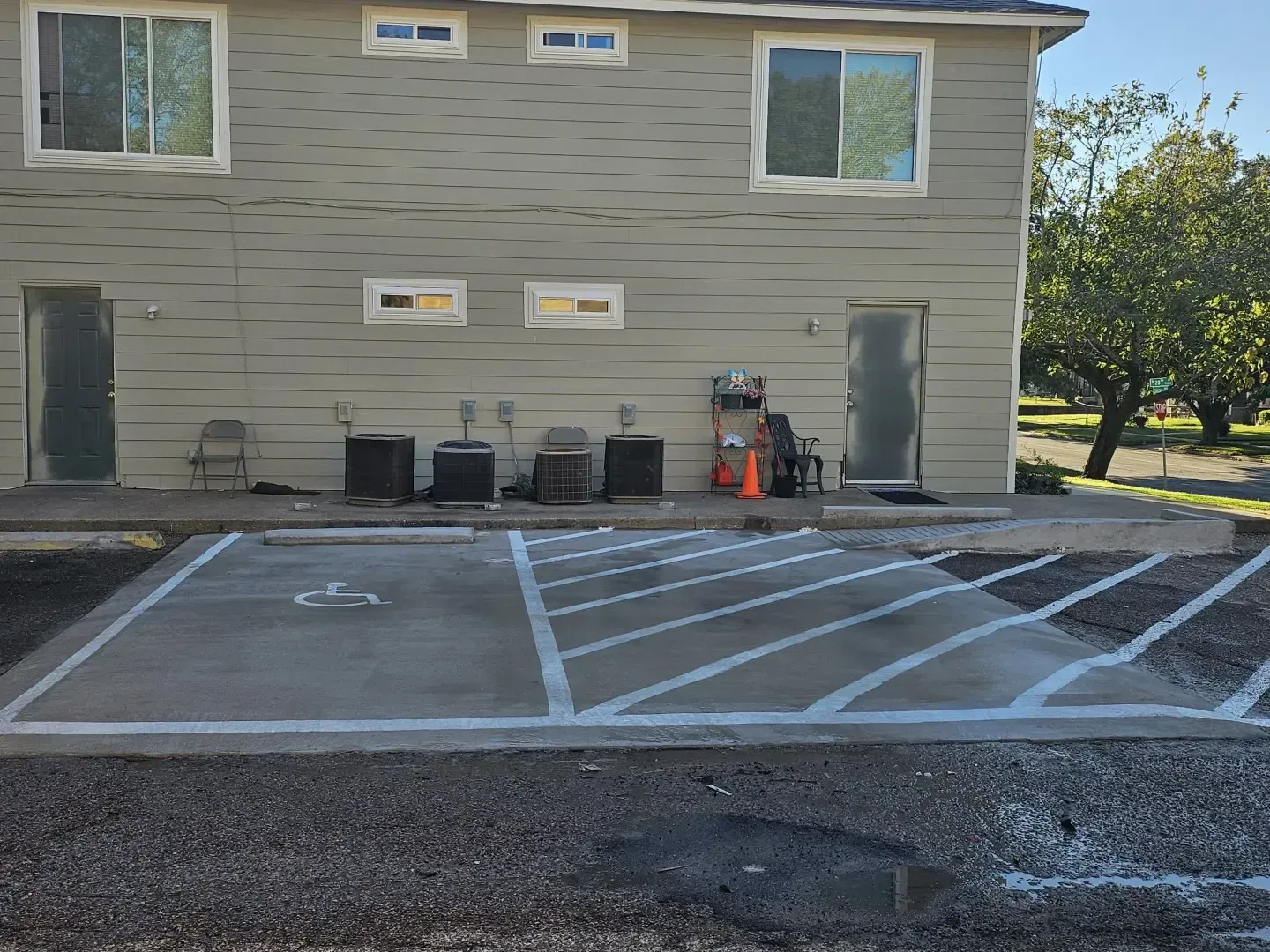 Accessible parking spaces marked in front of a beige building with two chairs and utility items against the wall